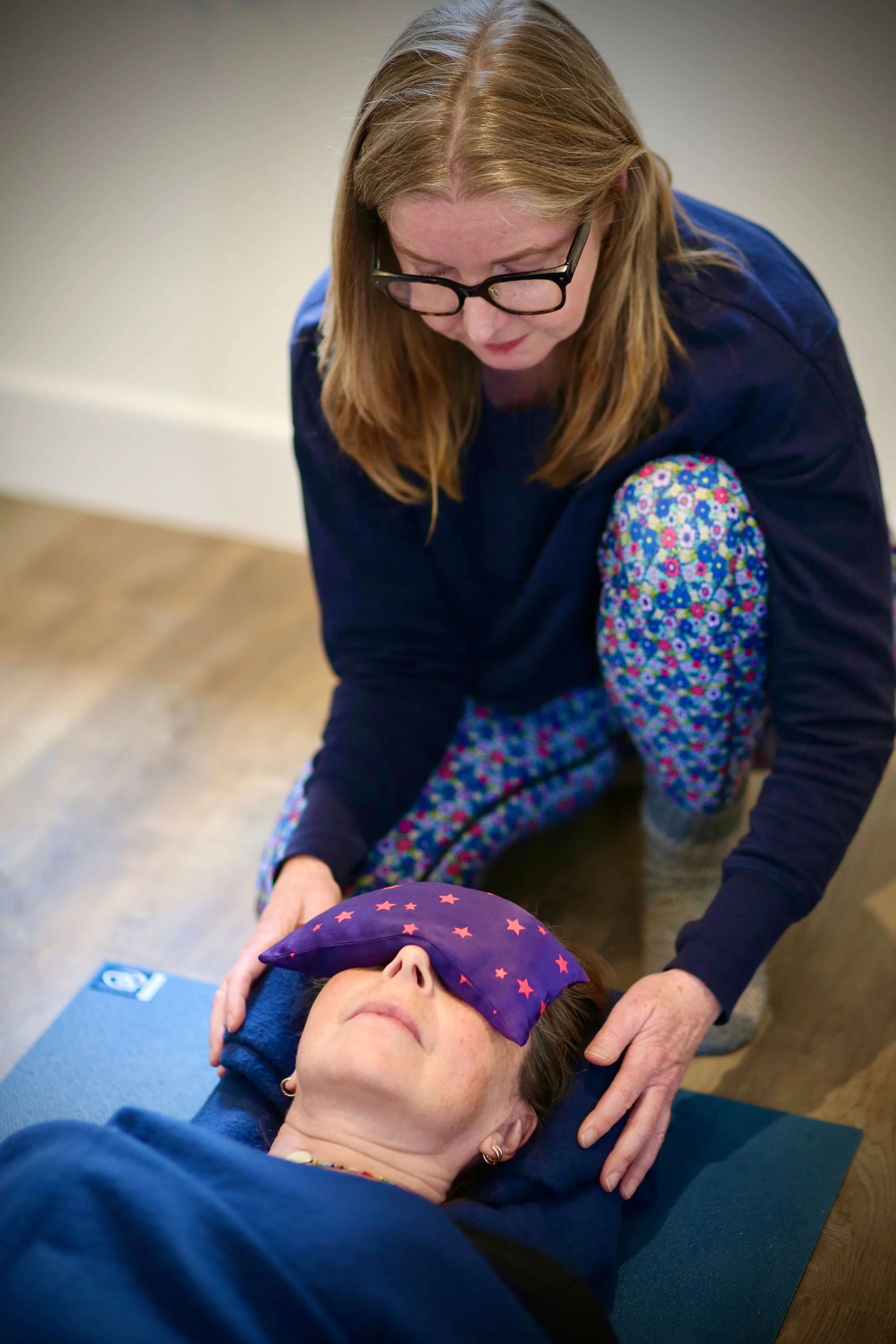 Catriona of Little Barn Yoga with long hair and glasses providing a Reiki session to a woman lying on a yoga mat, wearing a sleeping mask with stars, inside a room.