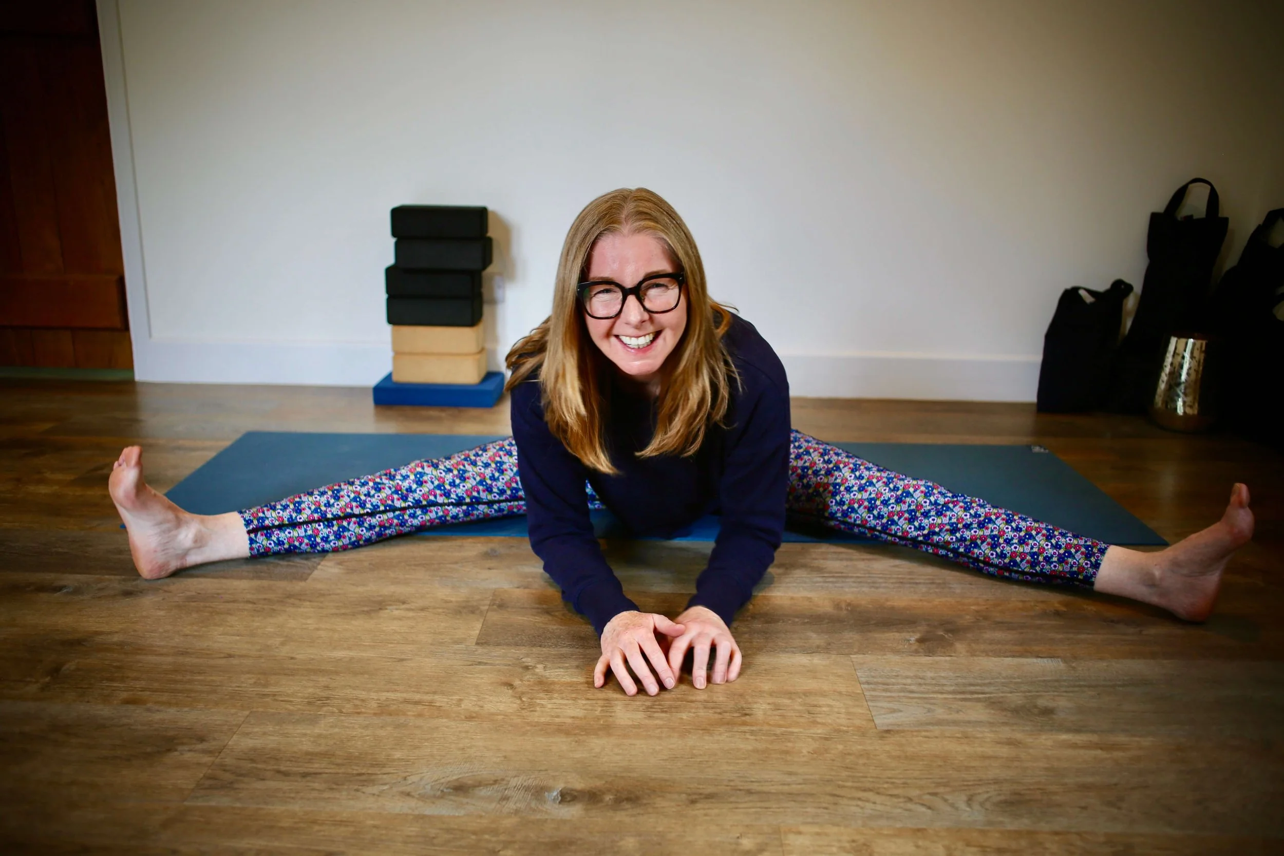 Catriona of Little Barn Yoga smiling in a yoga pose on a wooden floor, doing a full split with hands on the ground, in a room with a plain white wall, black and beige yoga blocks, a black bag, and a water bottle in the background.