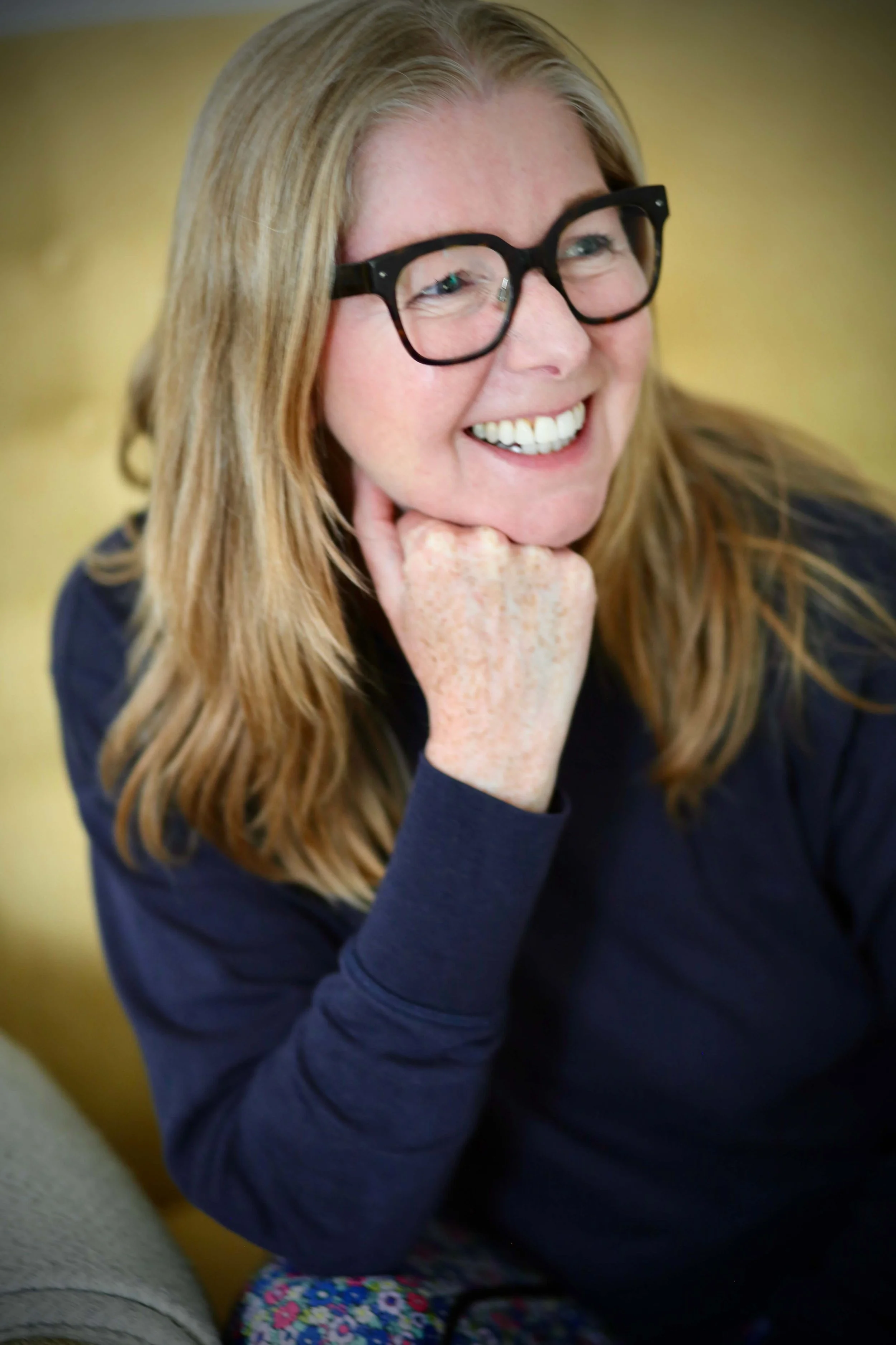 Catriona of Little Barn Yoga wearing black glasses and a dark blue shirt, smiling and resting her chin on her hand.