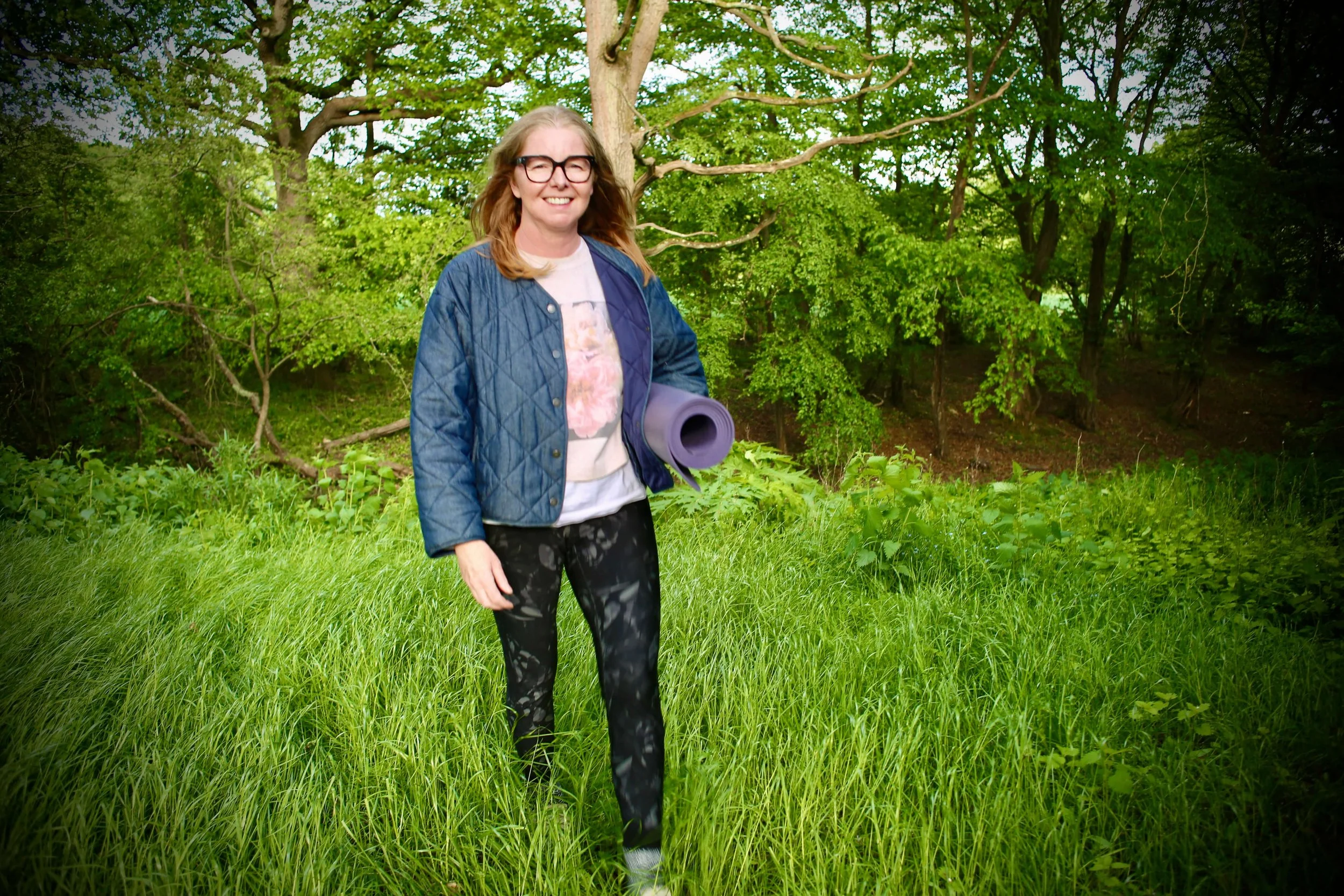 Catriona of Little Barn Yoga smiling outdoors holding a yoga mat, surrounded by green trees and grass.