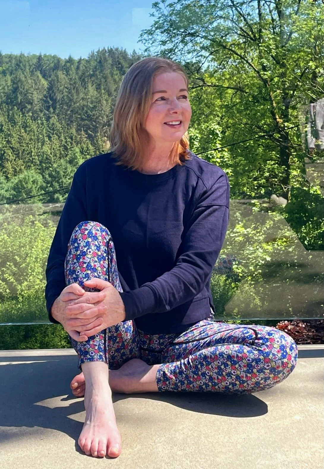 Catriona of Little Barn Yoga with shoulder-length hair is sitting cross-legged outdoors on a surface, with greenery and trees in the background under a clear blue sky. She is smiling and wearing a dark top and colorful, floral patterned pants.