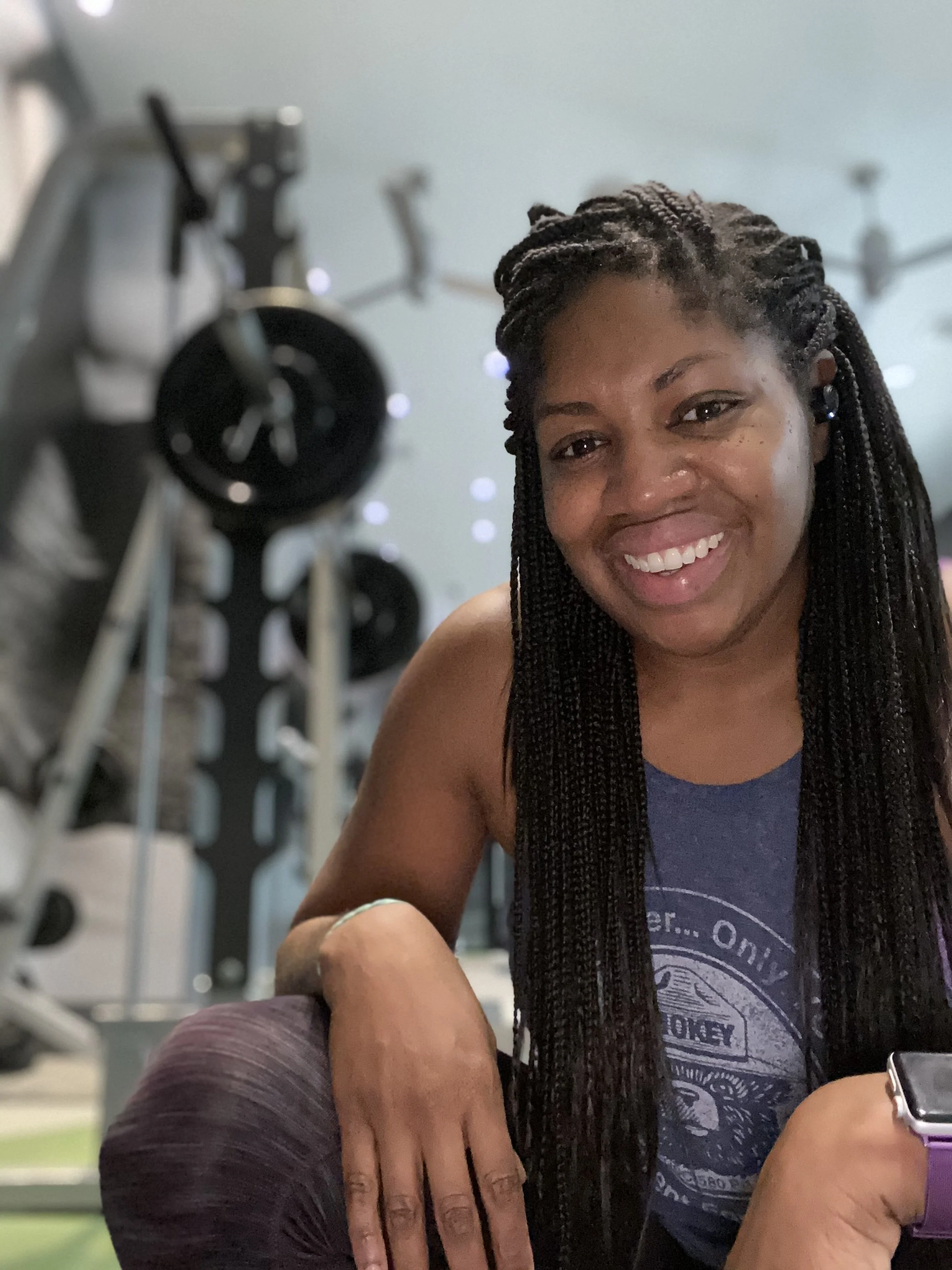 A woman with long braided hair smiling at the camera, seated with her arm resting on a surface, in a gym or fitness center with equipment in the background.