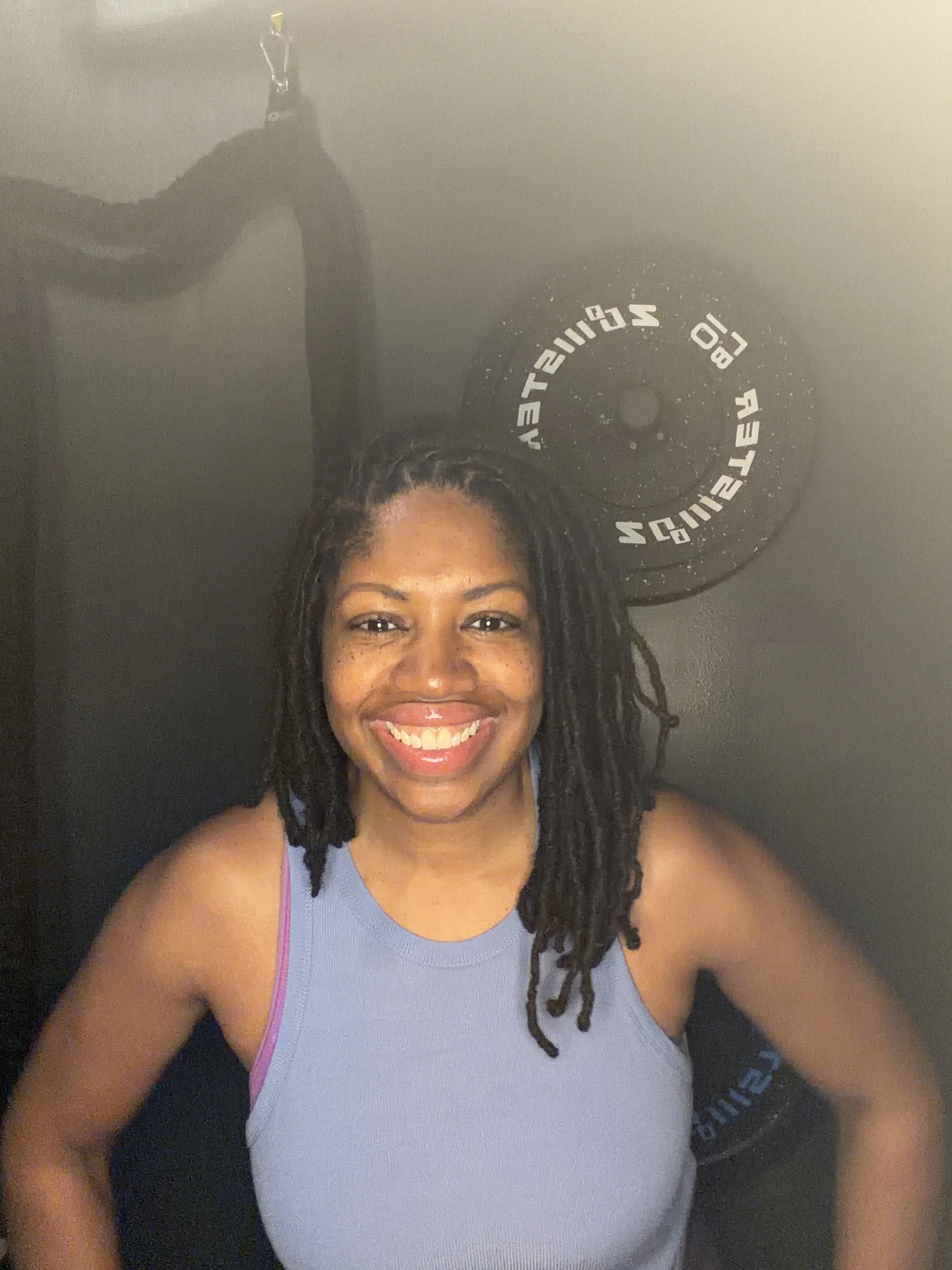 A woman smiling, wearing a light purple athletic top and black backpack, with a clock on the wall behind her.