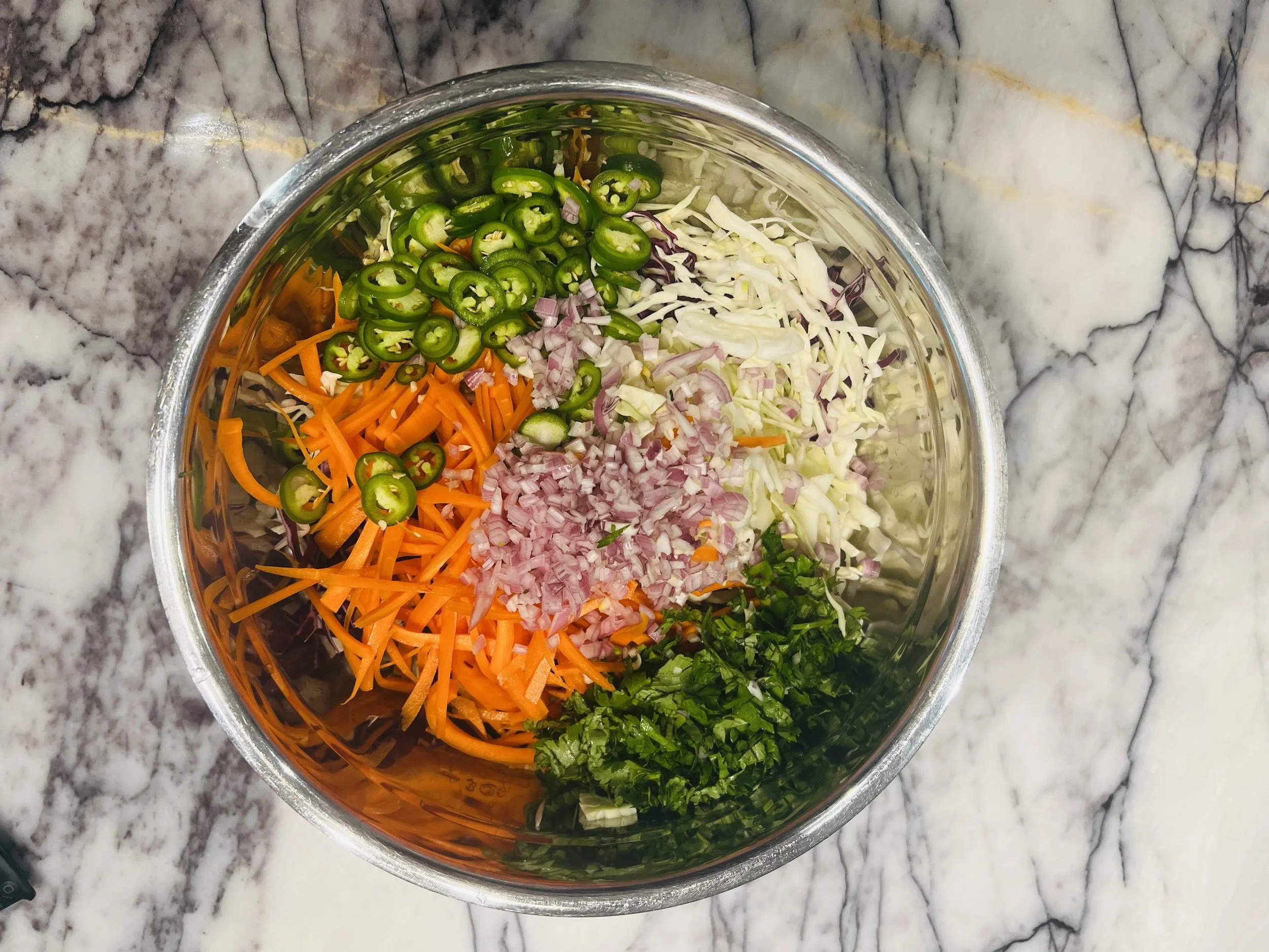 A glass bowl filled with chopped vegetables including green chili peppers, shredded cabbage, shredded carrots, chopped red onion, and chopped cilantro, placed on a marble countertop.