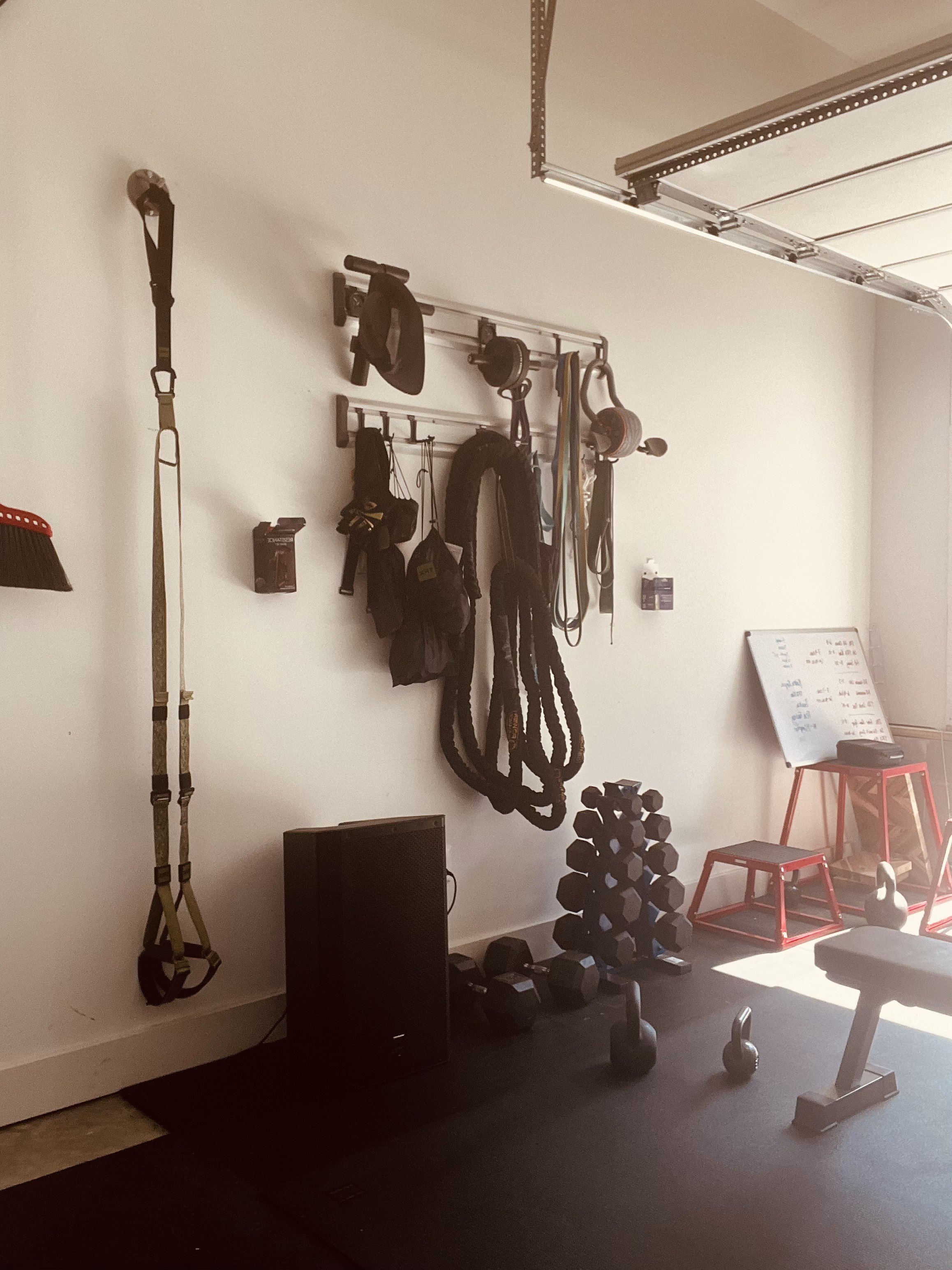 Home gym with wall-mounted racks holding suspension straps, kettlebells, dumbbells, medicine balls, and a whiteboard.