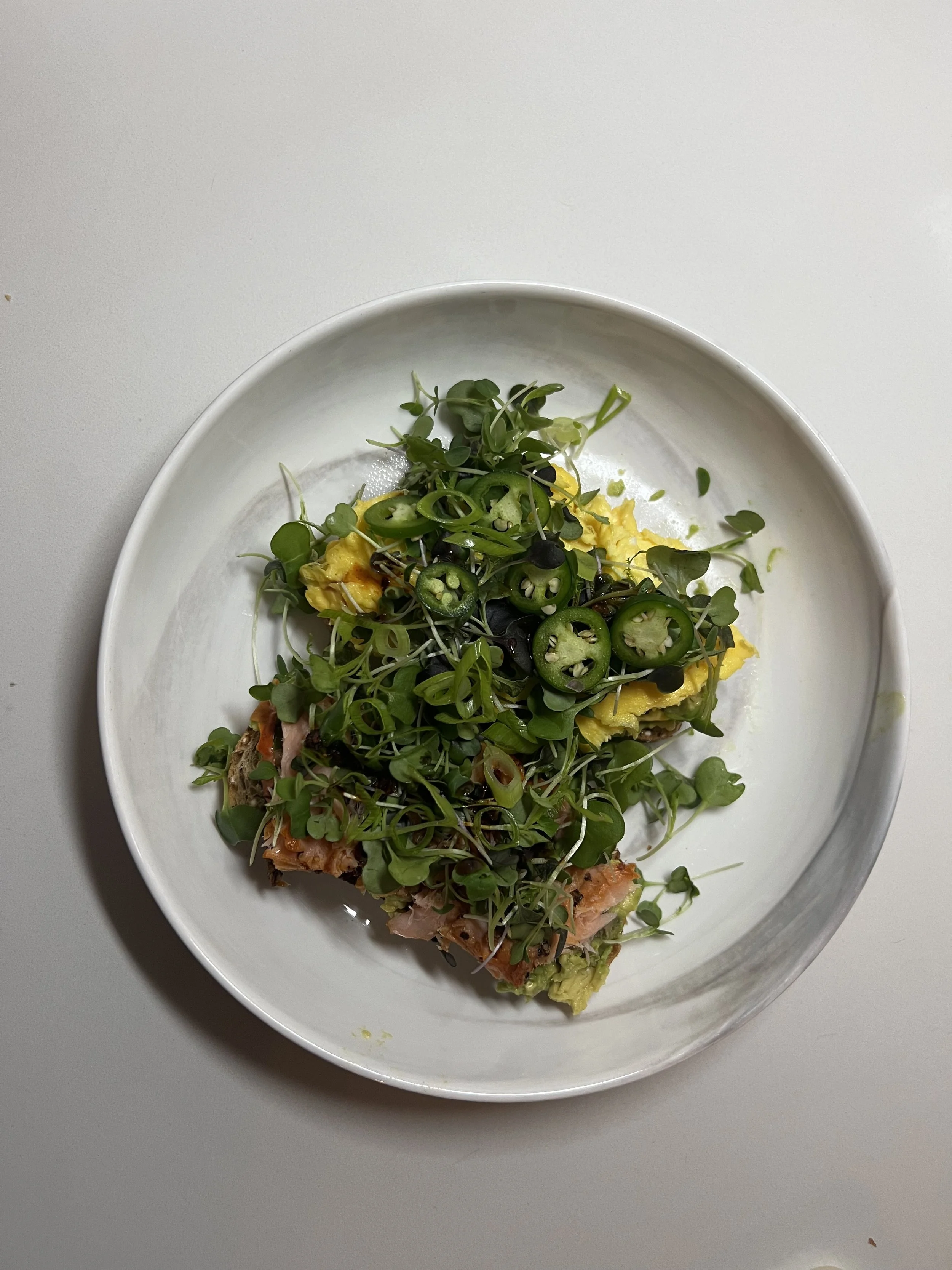 A white plate with scrambled eggs topped with microgreens, sliced green chili peppers, and other vegetables on a gray surface.