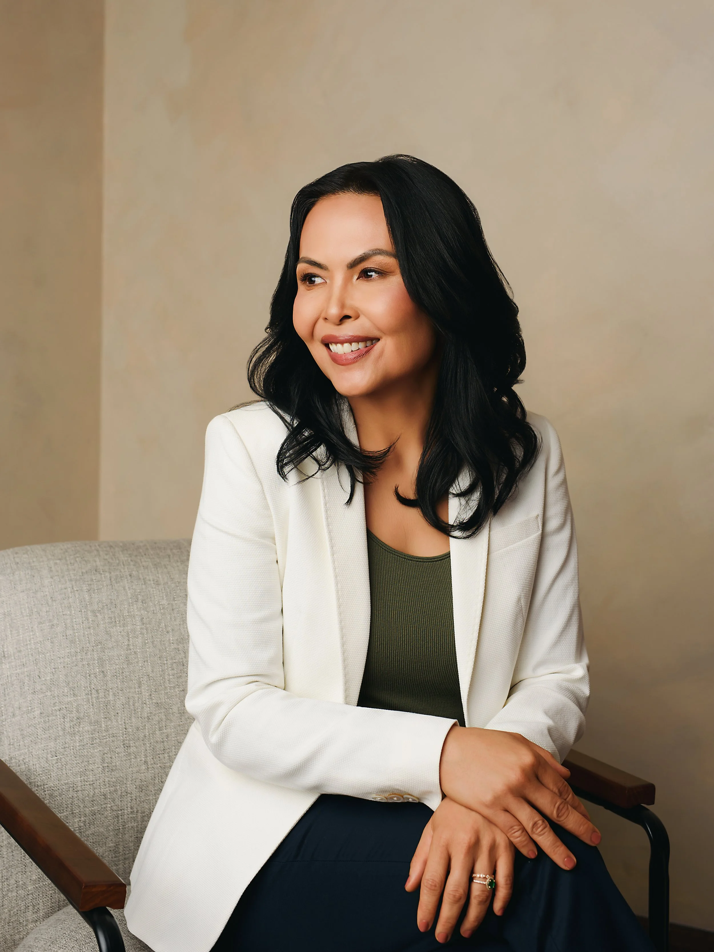 A woman with black hair and light skin tone, dressed in a white blazer and dark shirt, sitting on a beige chair with wooden armrests in a room with beige walls. She is smiling and looking to the right, with her right arm resting on her lap and her left hand on her right knee, wearing a ring with a green gemstone.