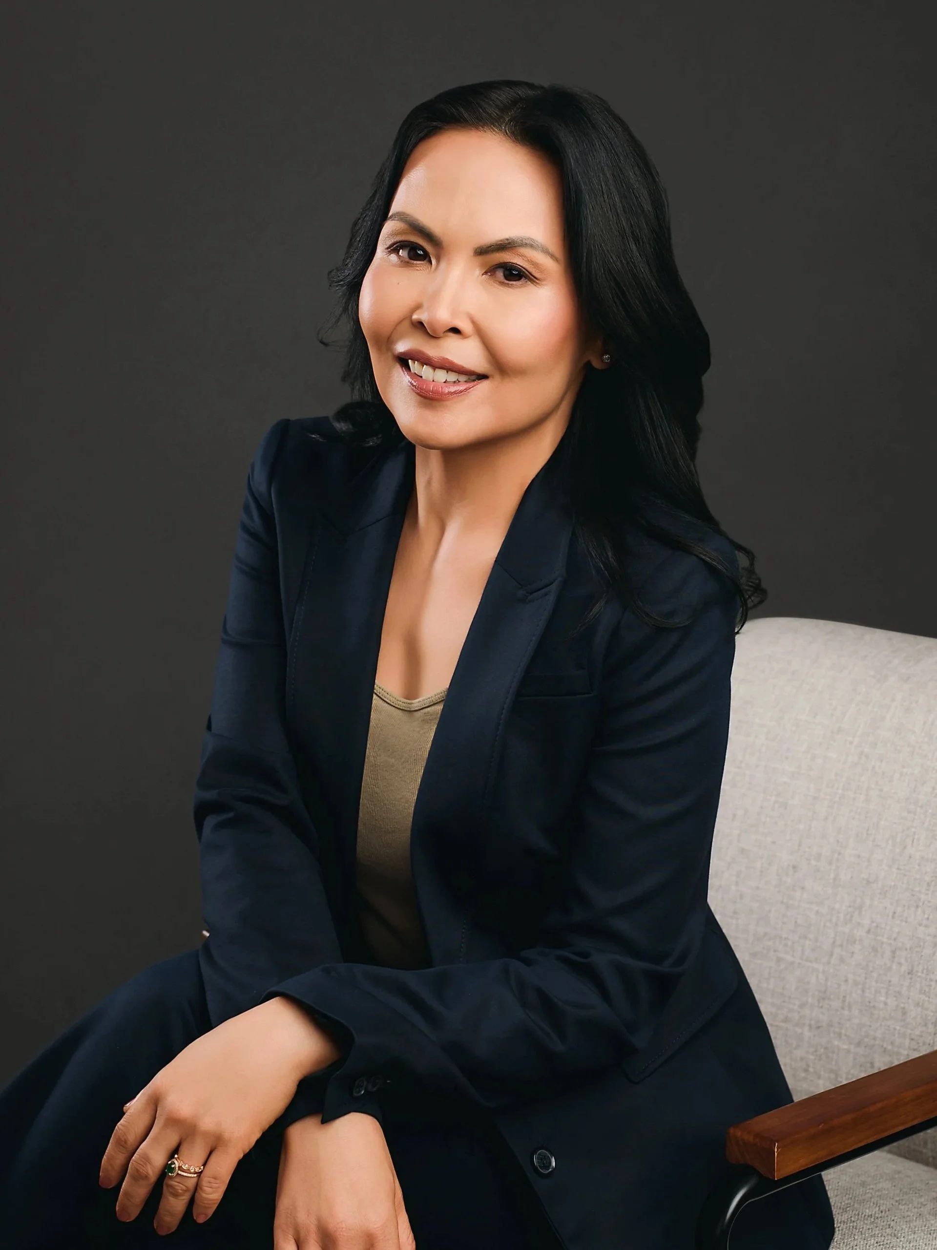 Portrait of a woman with black hair smiling, wearing a dark blue blazer and a beige top, sitting on a gray chair with a dark gray background.