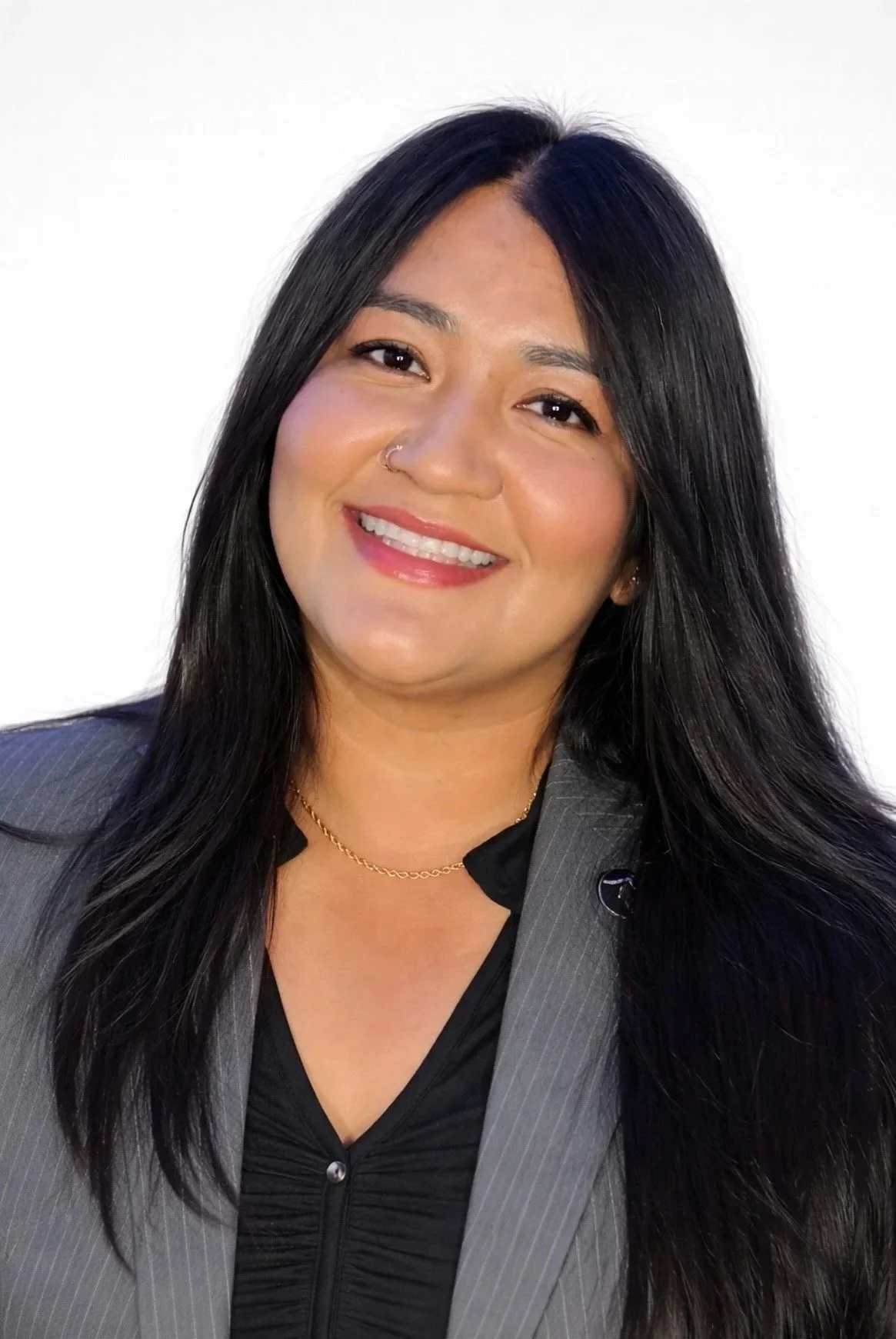 A woman with long black hair, wearing a gray blazer, black top, gold necklace, and a nose ring, smiling in front of a plain white background.