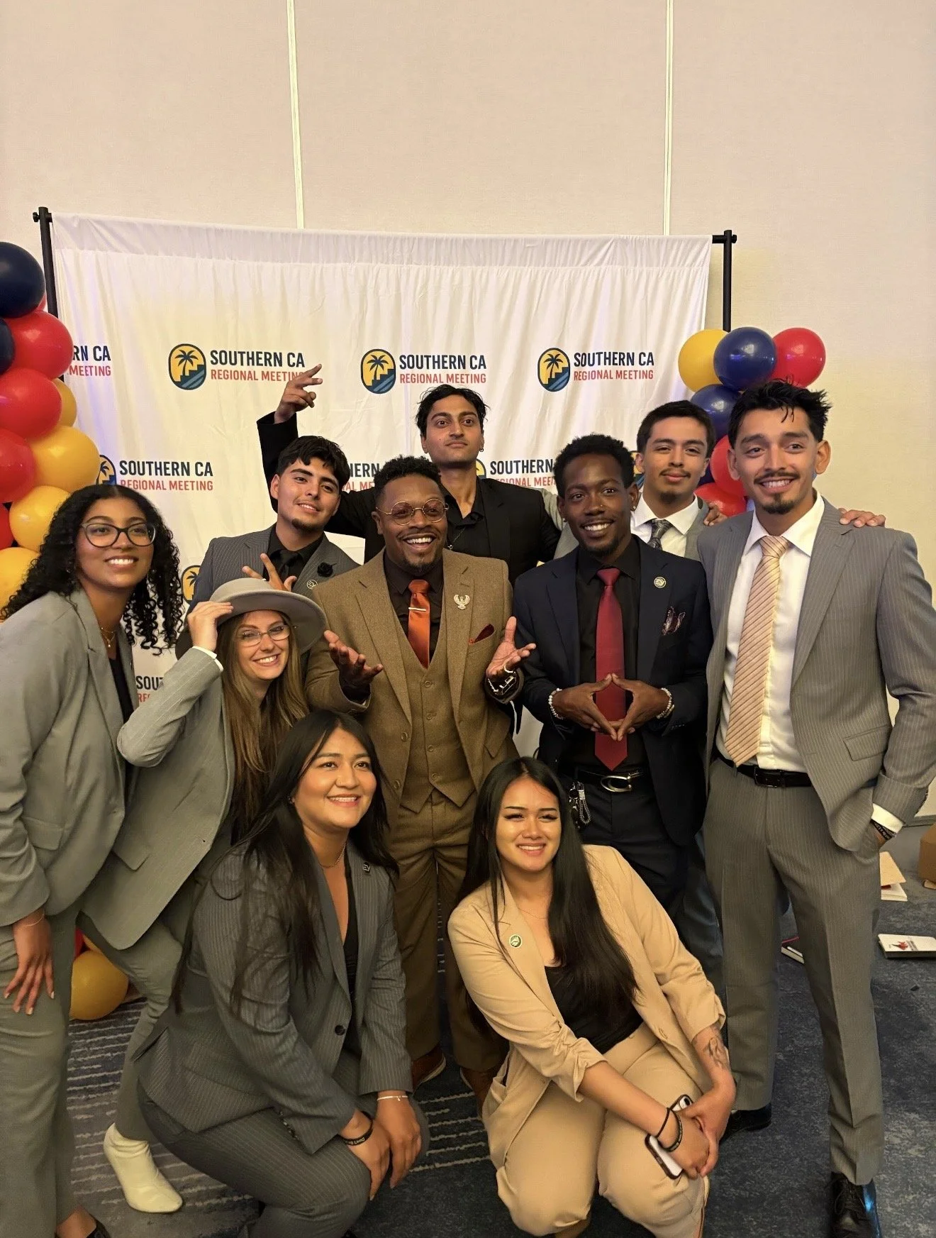 A group of diverse young adults posing together at a Southern California regional meeting, standing in front of a white backdrop with the event logo, surrounded by colorful balloons.