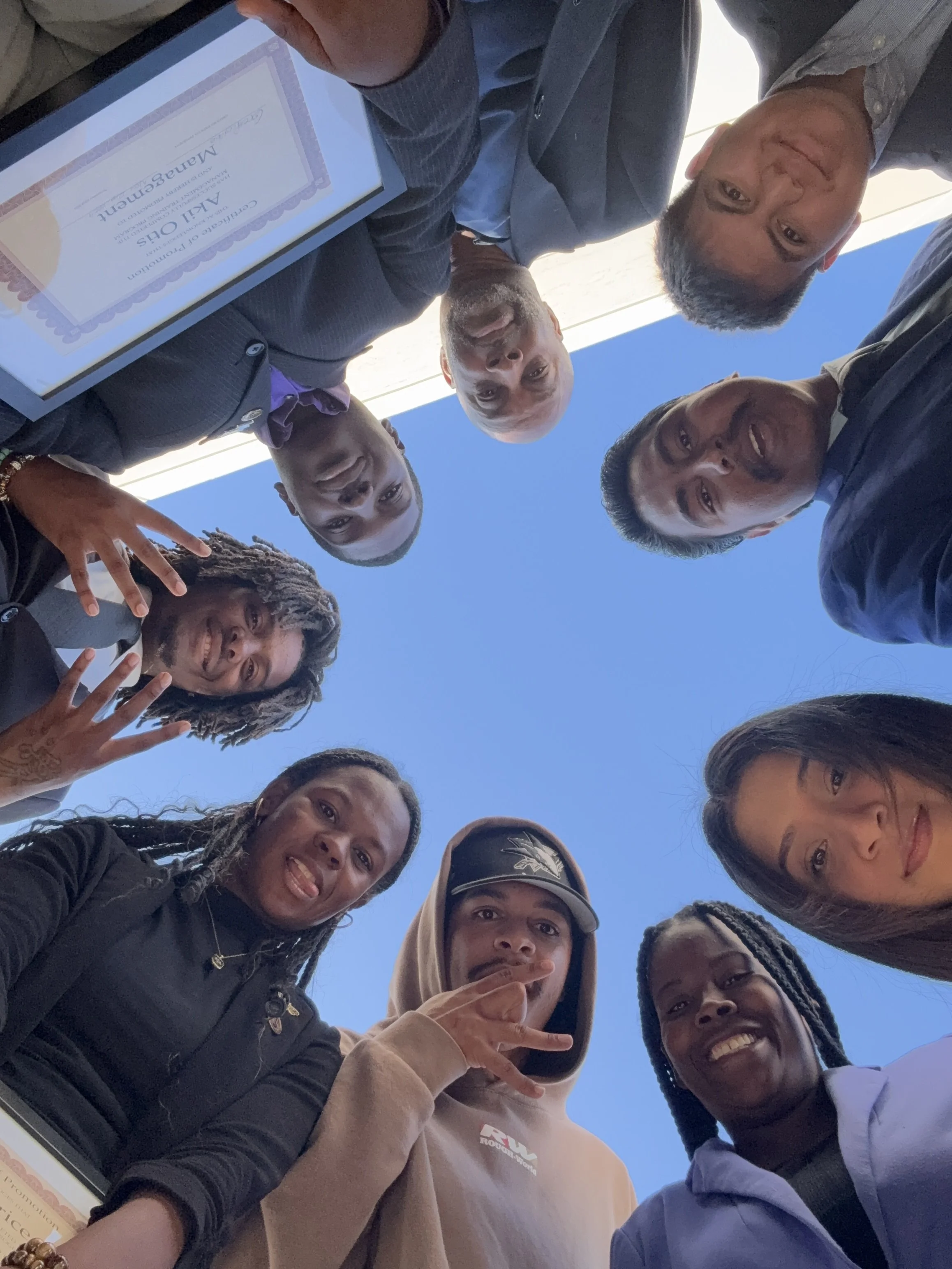 A group of diverse people smiling and looking down at the camera, forming a circle outdoors against a clear blue sky. One person is holding a framed certificate.