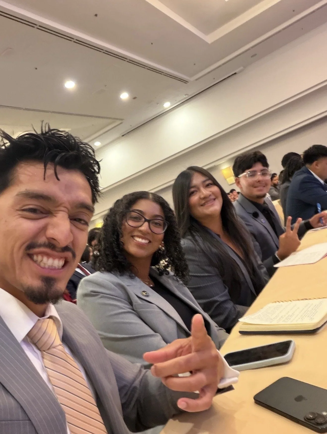 Group of four people sitting at a conference table, smiling and giving thumbs up in a conference room filled with other attendees.