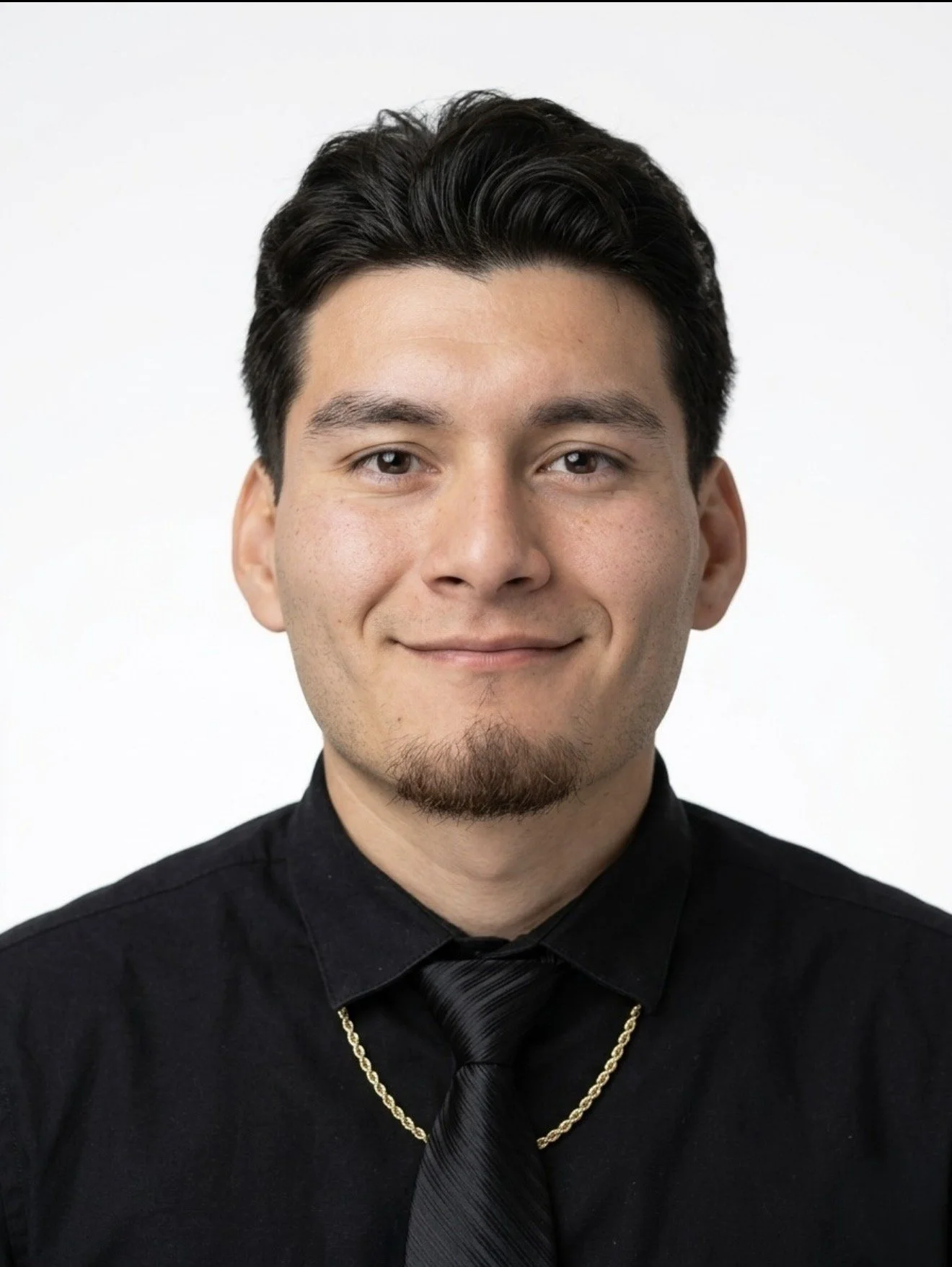 A young man with dark hair styled back, wearing a black dress shirt, a black tie, a gold chain, and a slight smile, posed against a plain white background.