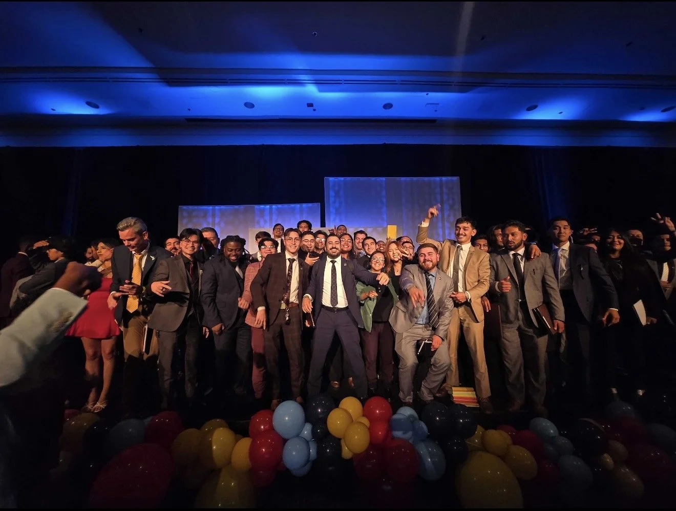 Group of people in formal attire on stage at a celebration or event, with balloons at their feet and dim lighting overhead.