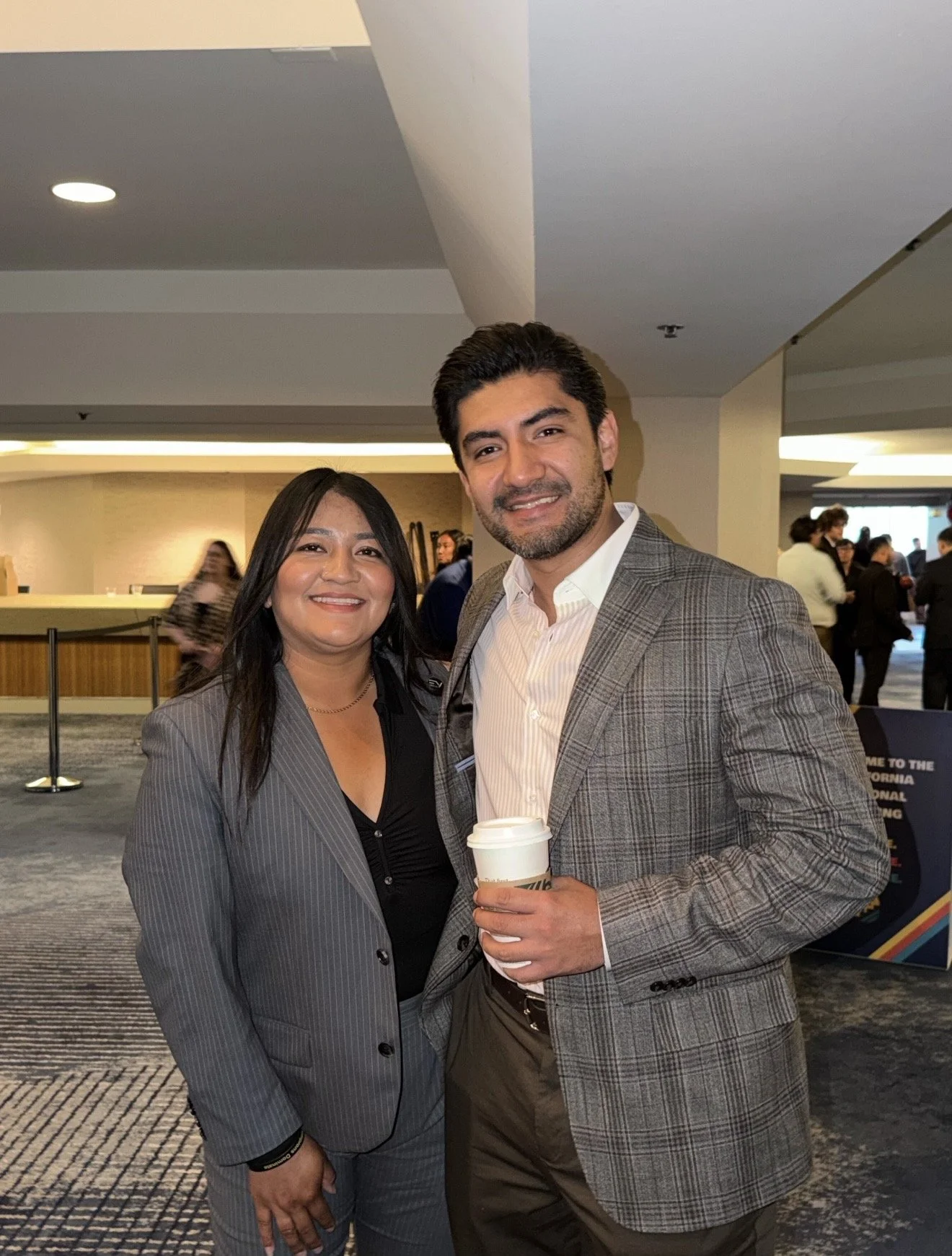 A man and woman posing together indoors at a social event, both smiling. The man is holding a coffee cup. Background shows other people and a sign.