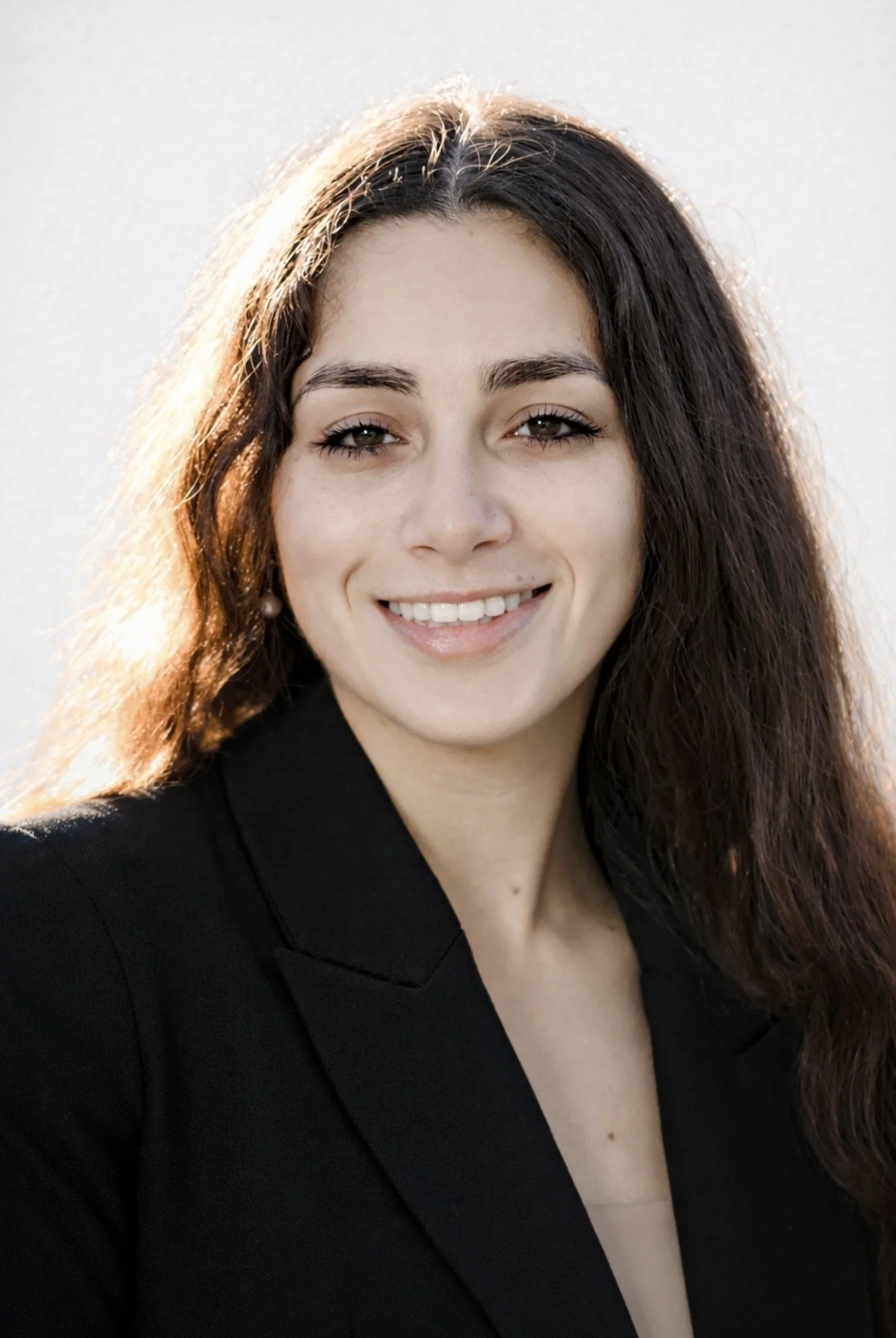 Close-up of a smiling woman with long brown hair wearing a black blazer and pearl earrings, outdoors with sunlight behind her.