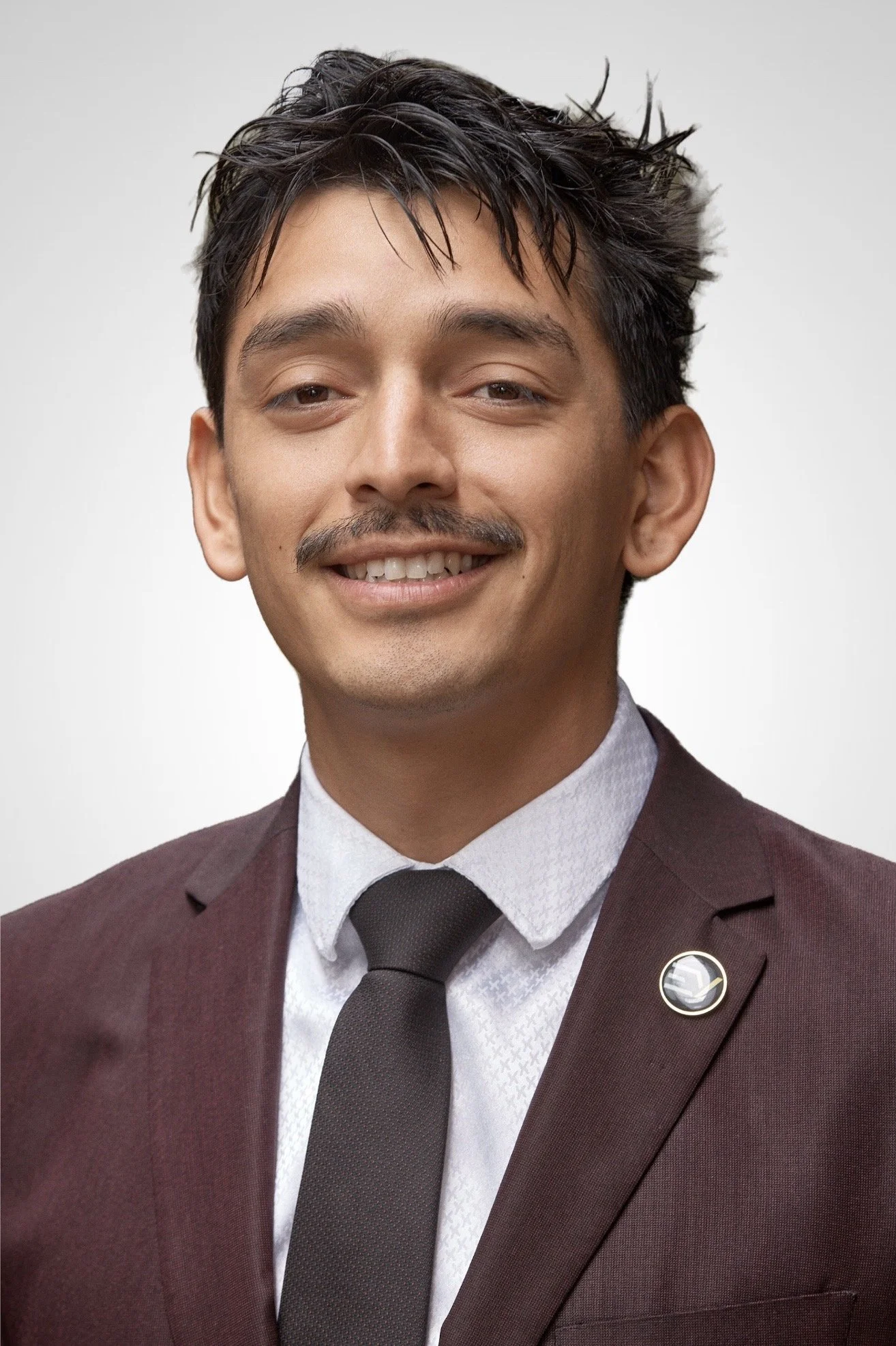 A man in a burgundy suit with a pin, white shirt with a textured pattern, and black tie, smiling against a plain background.