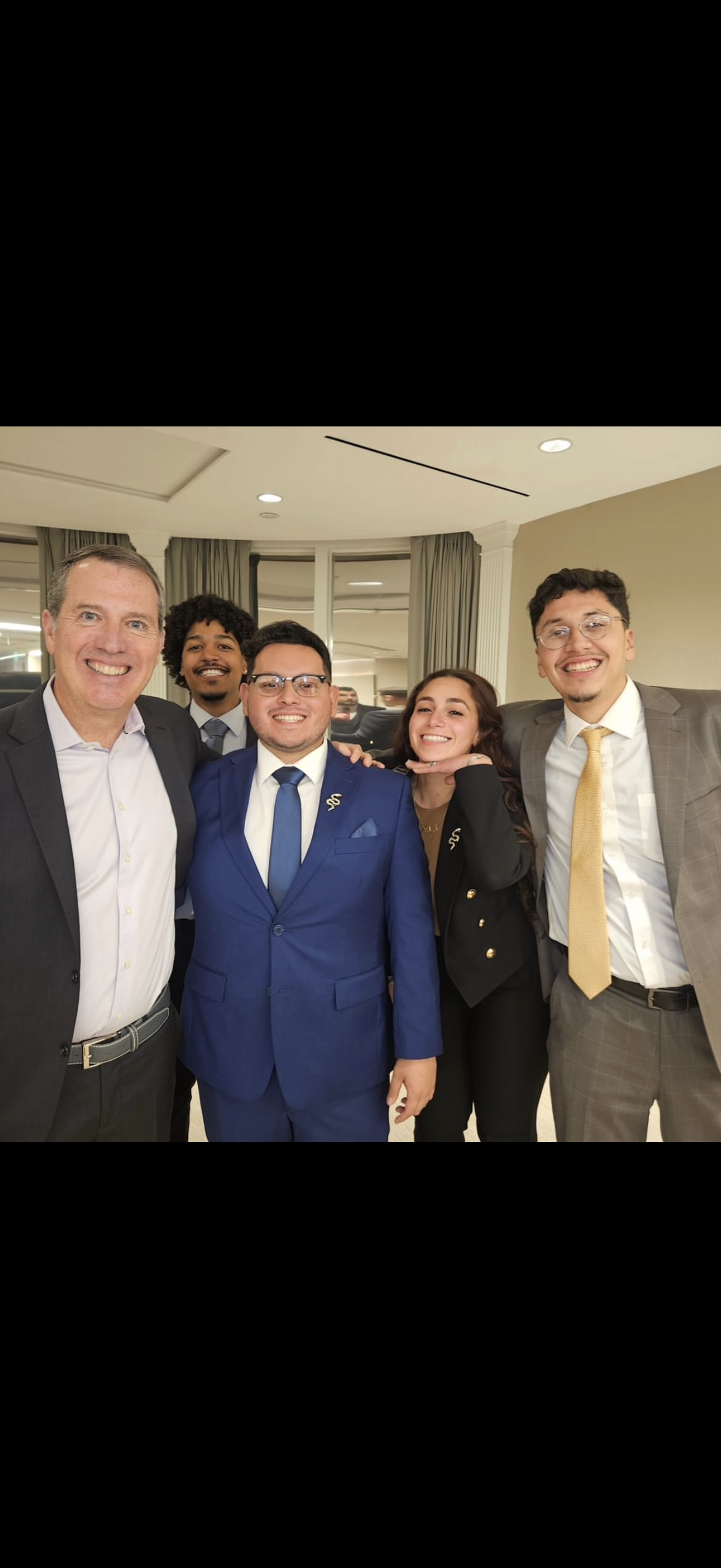 Group of five people dressed in formal attire, smiling, indoors at a celebration or event.