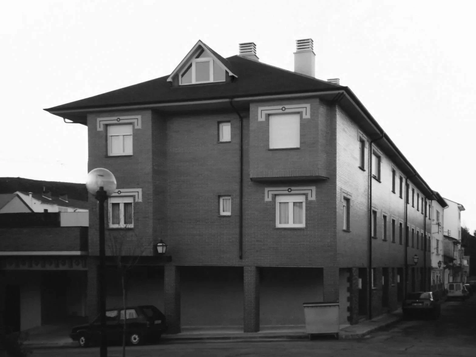 Edificio de tres pisos con ventanas con cortinas y chimeneas en el tejado, en una calle con lampas y coches estacionados.