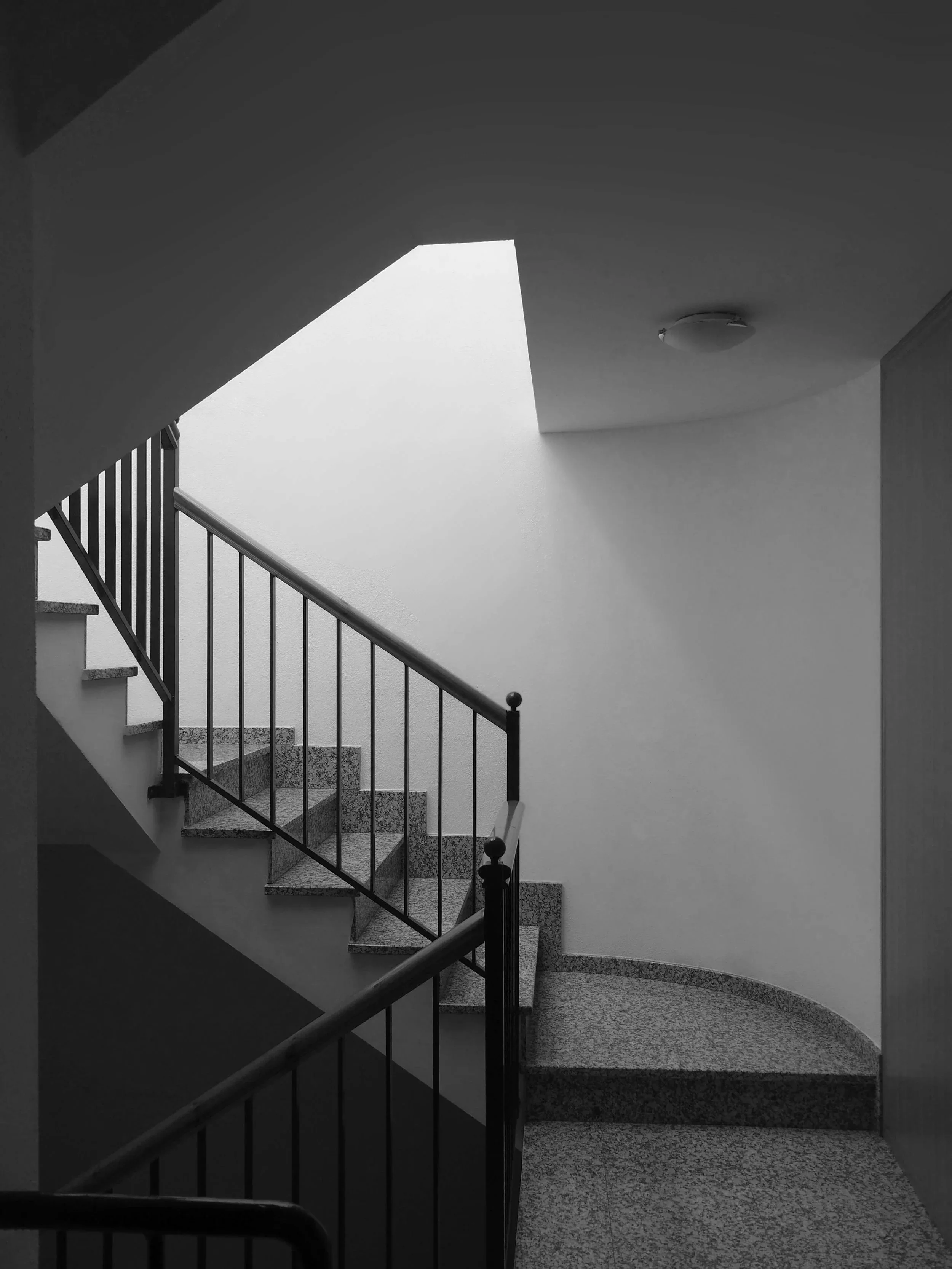 Escalera interior de edificio con barandillas metálicas y paredes blancas, vista desde la parte inferior hacia arriba, iluminada por luz natural.