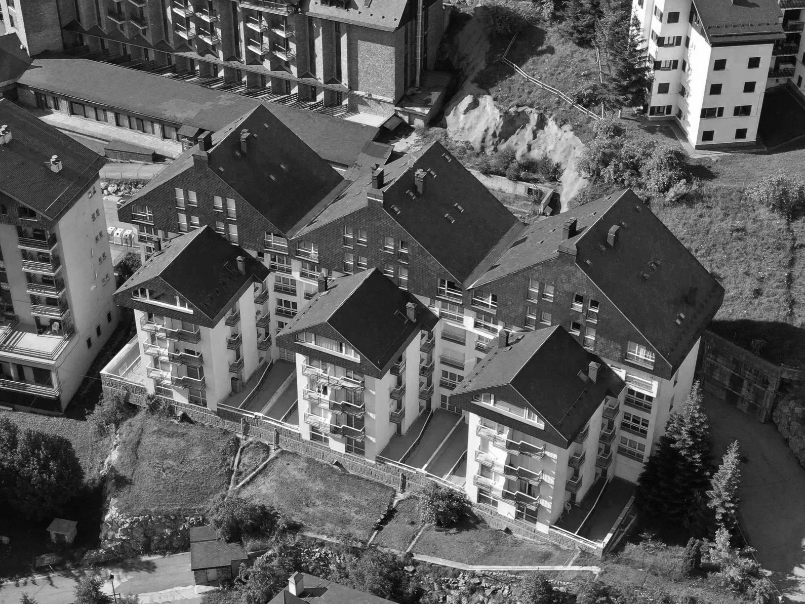 Vista aérea en blanco y negro de edificios residenciales, algunas con balcones, y terrenos verdes y de tierra en el entorno.