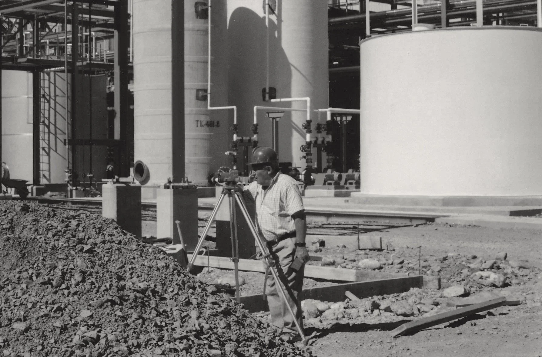 Trabajador en una obra industrial con equipo de medición y casco en un sitio de construcción con tanques grandes y tuberías.