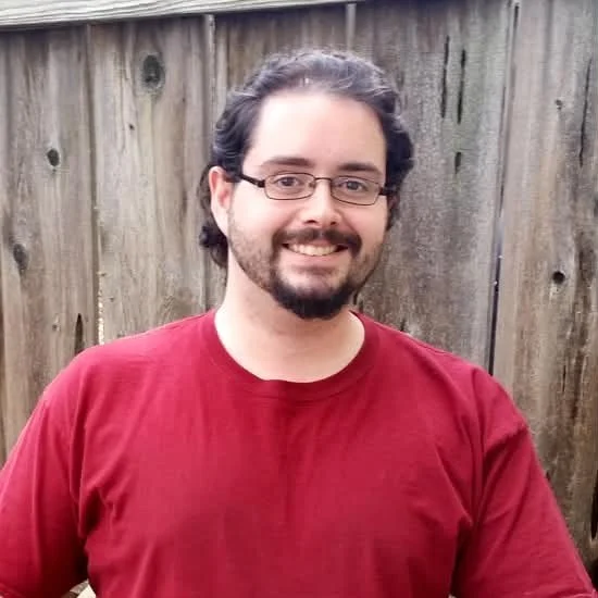 Man with glasses and beard wearing a red shirt, smiling outdoors in front of wooden fence.