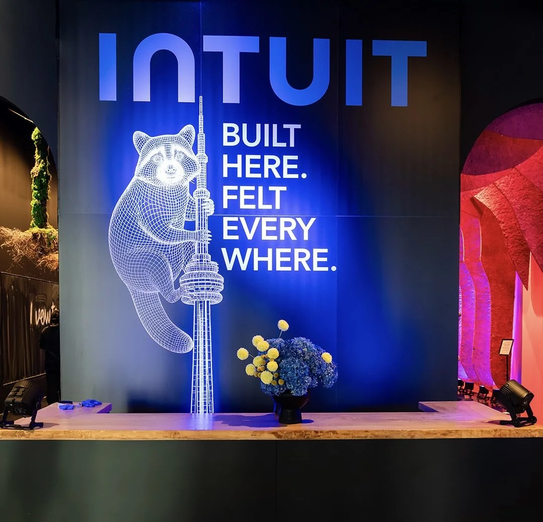 Large illuminated sign with the word 'INTUIT' and a digital wireframe of a raccoon holding a tower, with the text 'Built here. Felt everywhere.' and a flower arrangement in front.