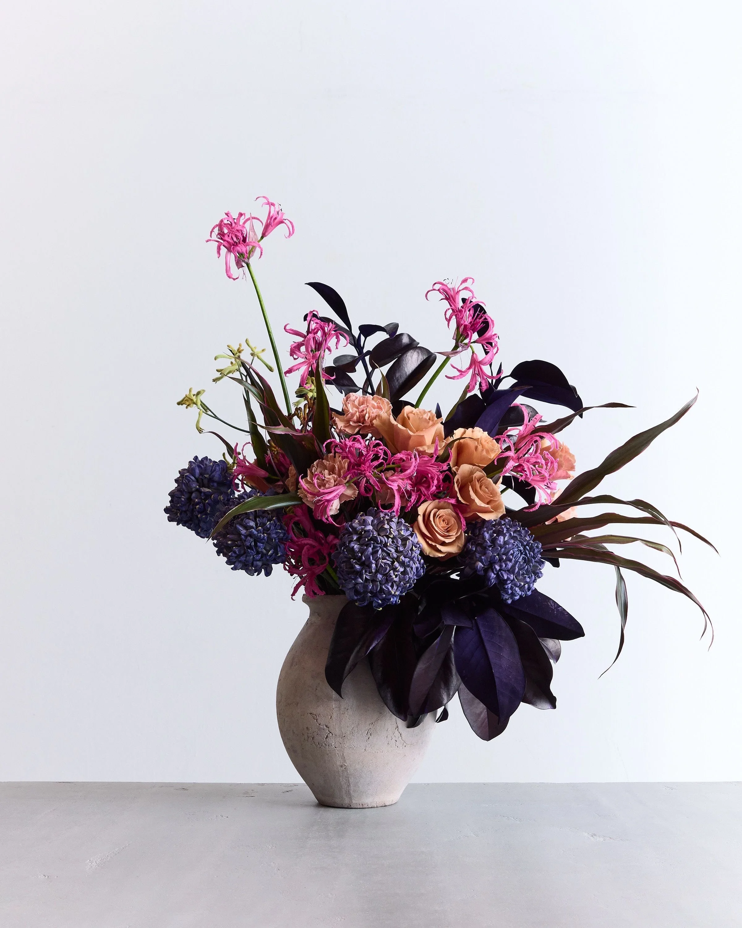 A minimalist floral arrangement in a textured white ceramic vase with purple, pink, and peach flowers and dark green and purple leaves, set against a plain white background.