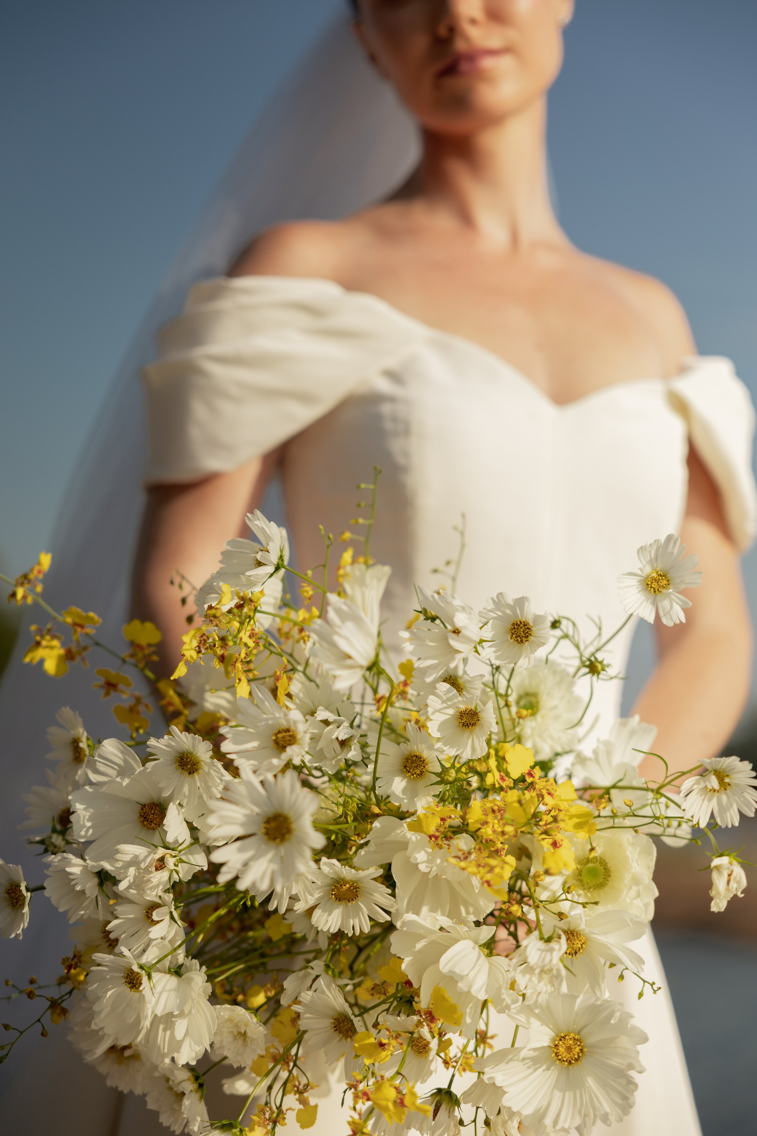 A woman in a white dress holding a large bouquet of white and yellow flowers. She is standing outdoors with a blue sky in the background, and a sheer veil drapes over her shoulder.