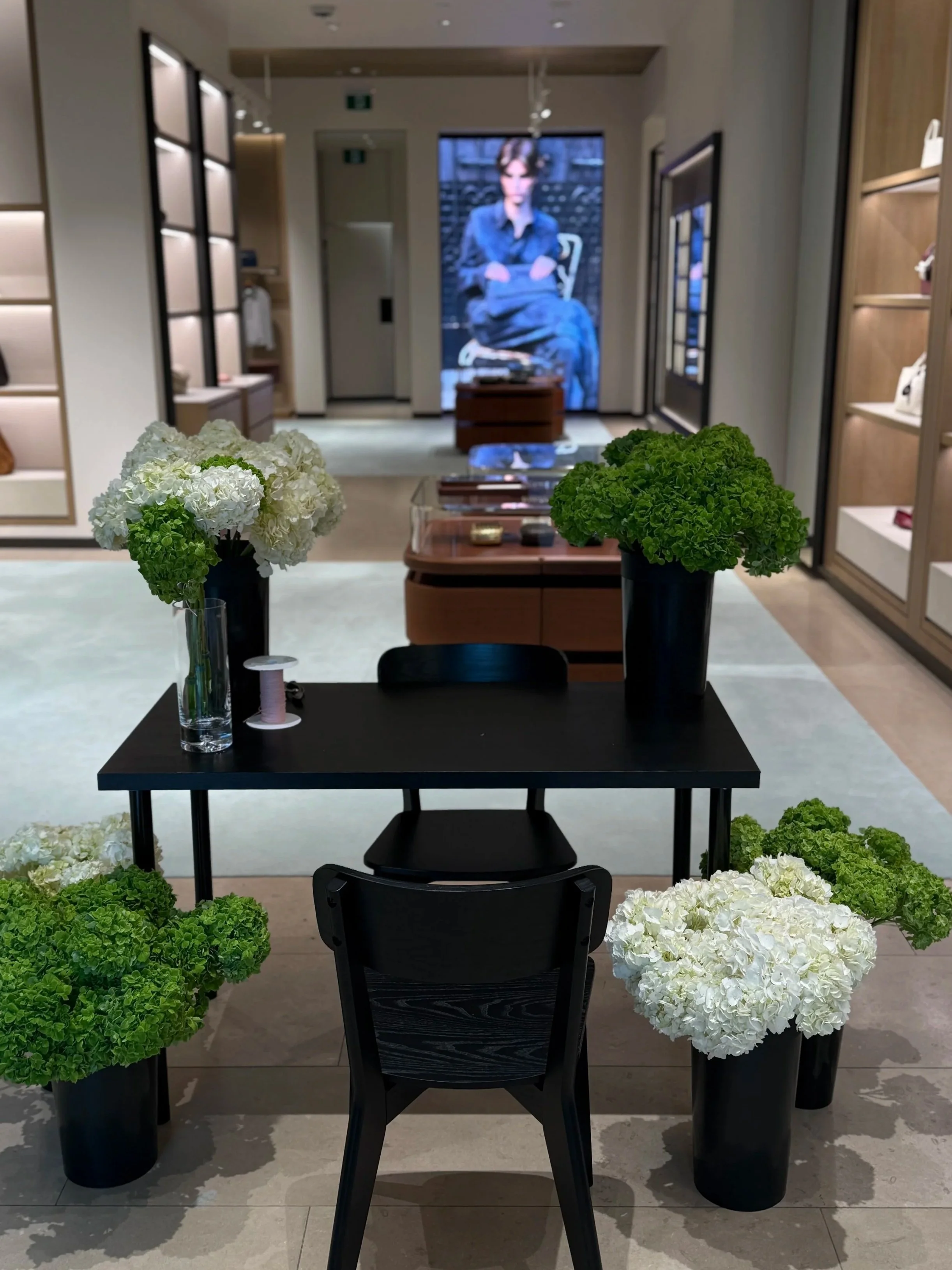 Indoor retail store display with a black table, green and white floral arrangements in black vases, and a seated black chair. Bottege Veneta, Toronto.