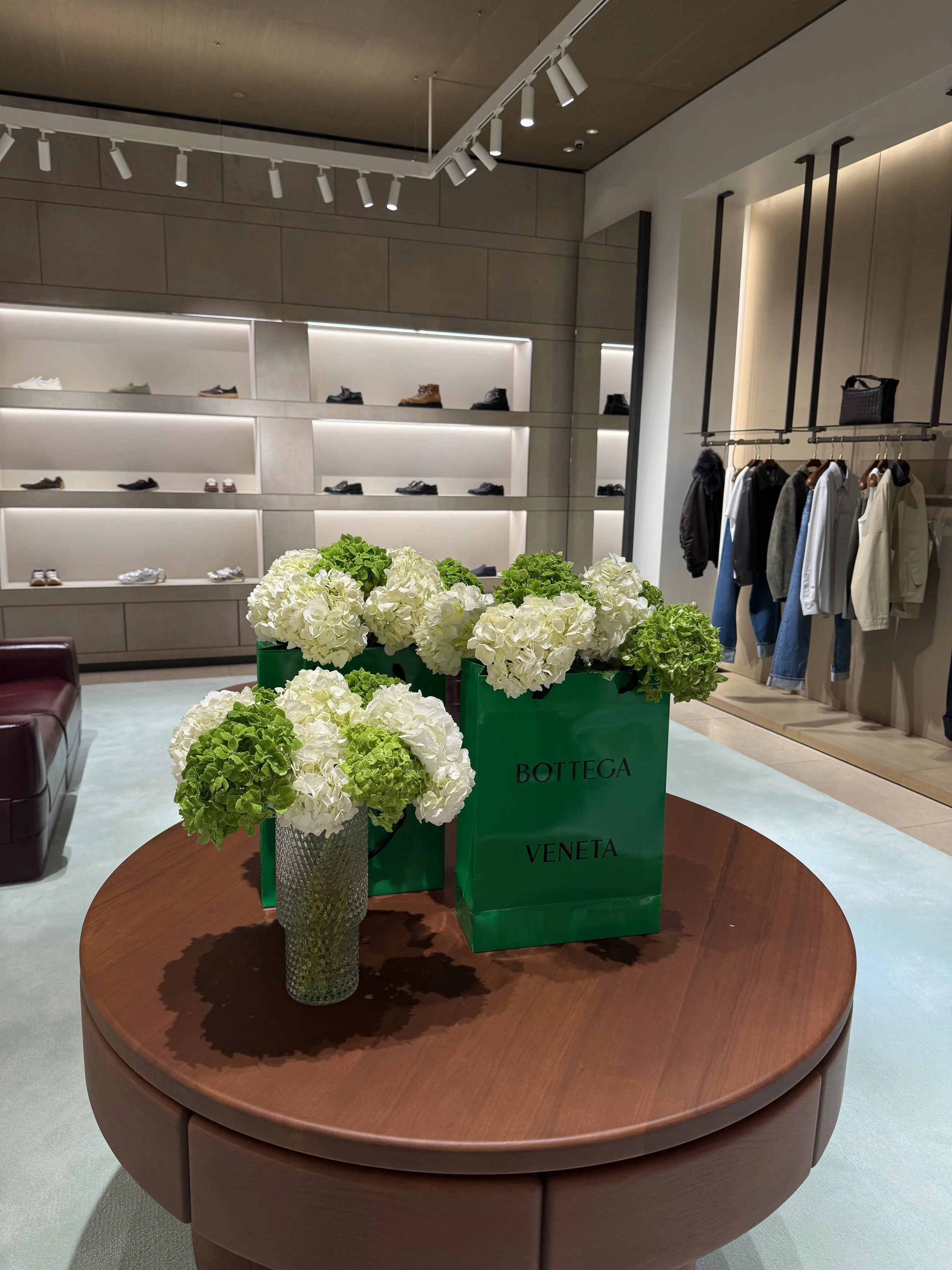A wooden round table with a vase of white and green hydrangea flowers and green Bottega Veneta shopping bags in a retail store.