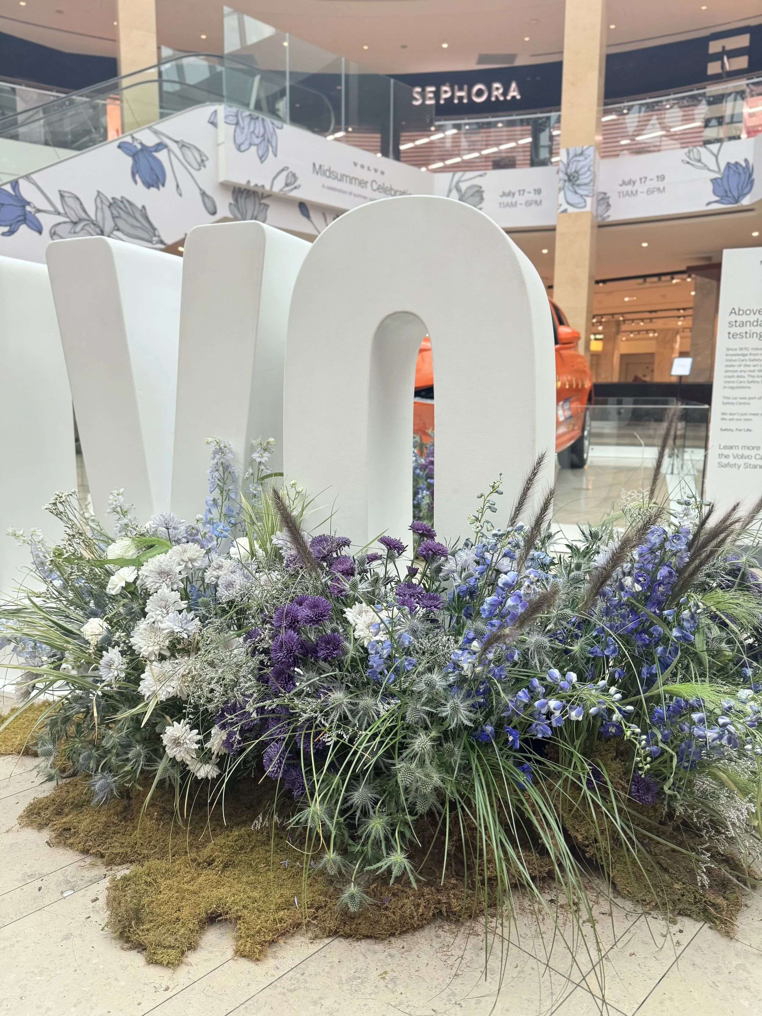 A large floral display with various purple, white, and blue flowers, green leaves, and grasses sits on a naturalistic base of moss in a shopping mall. Behind the display, there are multi-level shopping areas with stores, including a visible Sephora, 