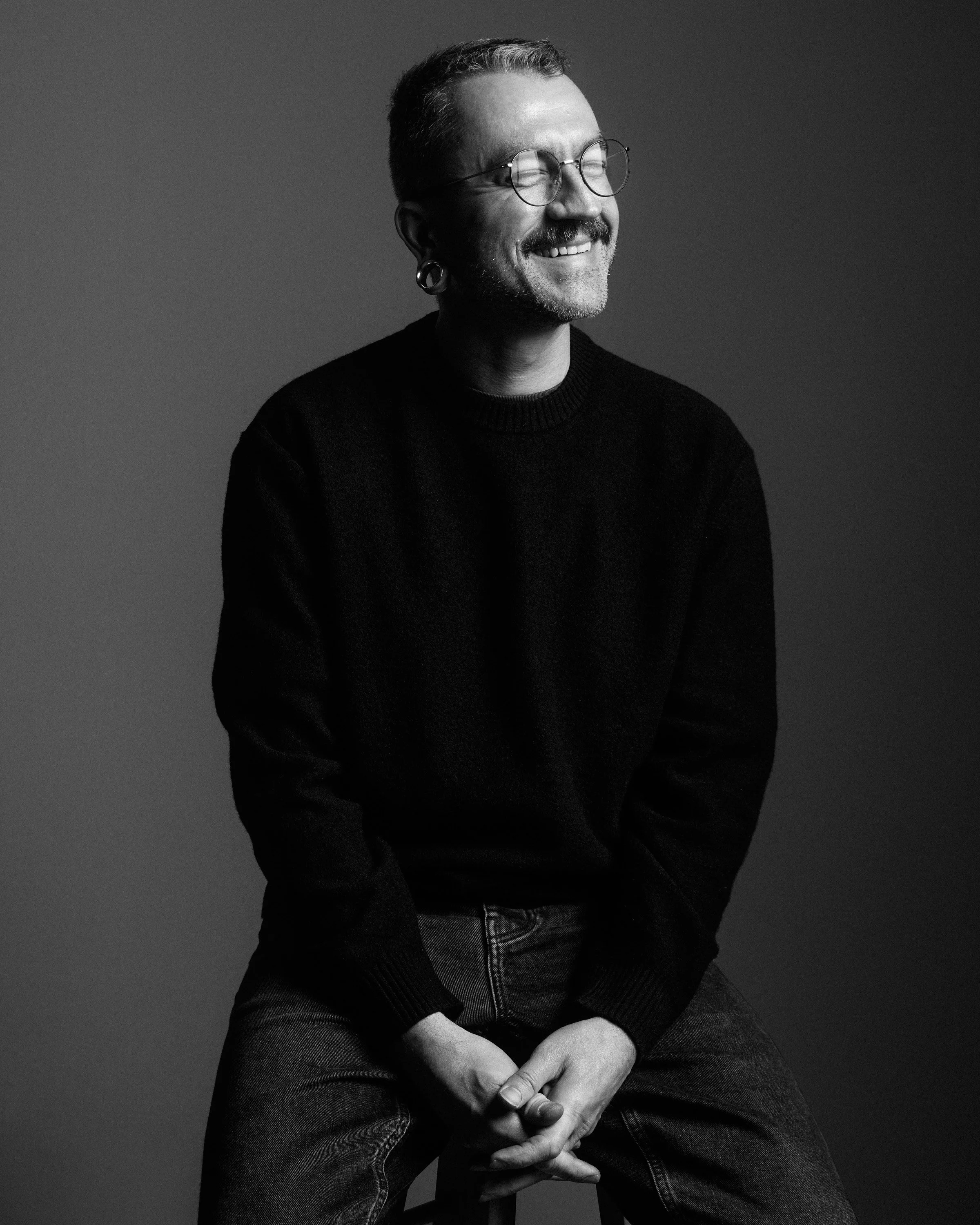 Black and white portrait of a smiling man with glasses and earrings, sitting with crossed hands, wearing a dark sweater and jeans.