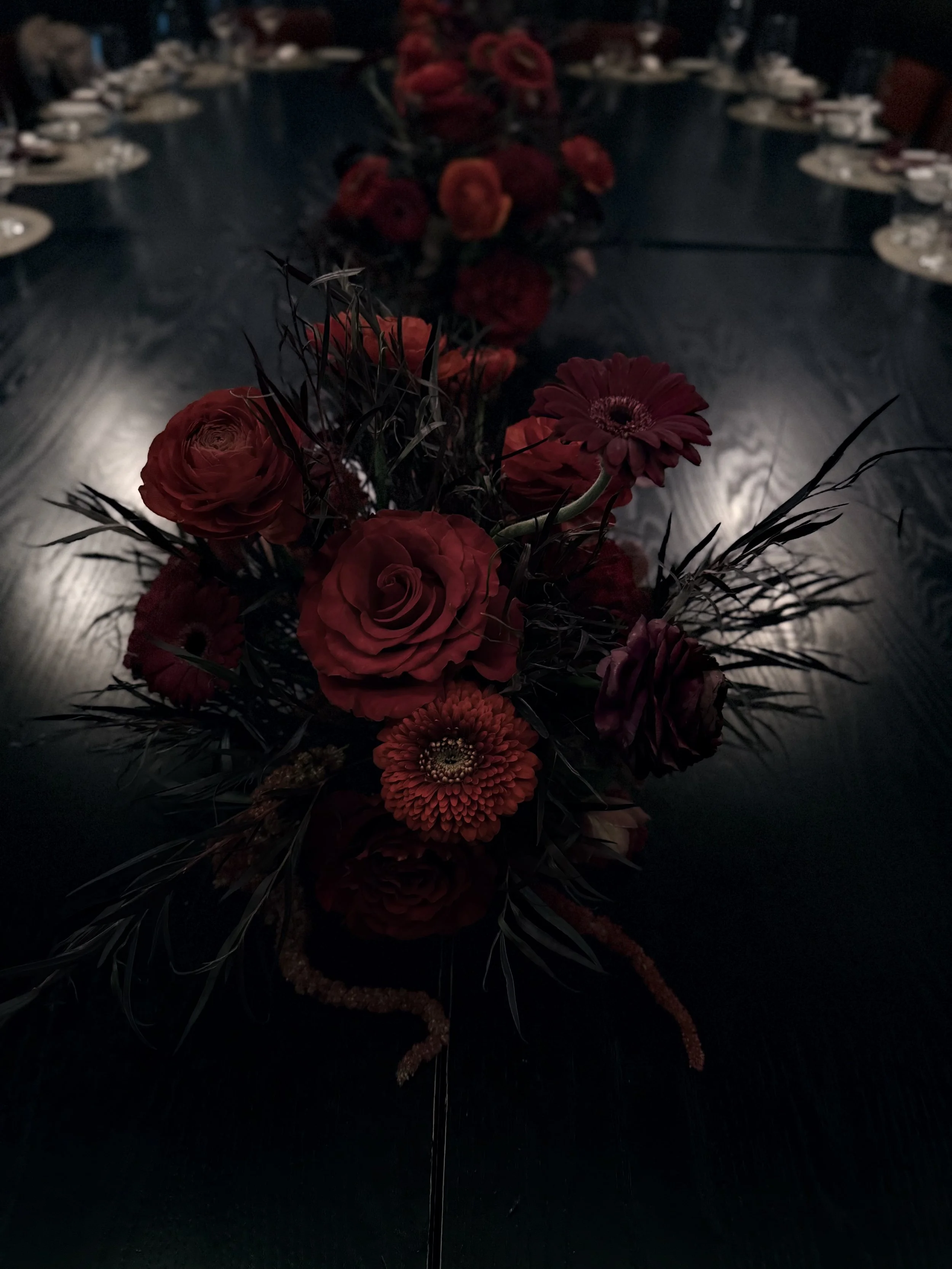 A bouquet of red and orange flowers, including roses, gerbera daisies, and ranunculus, with dark green foliage, arranged on a dark surface with a blurred background.