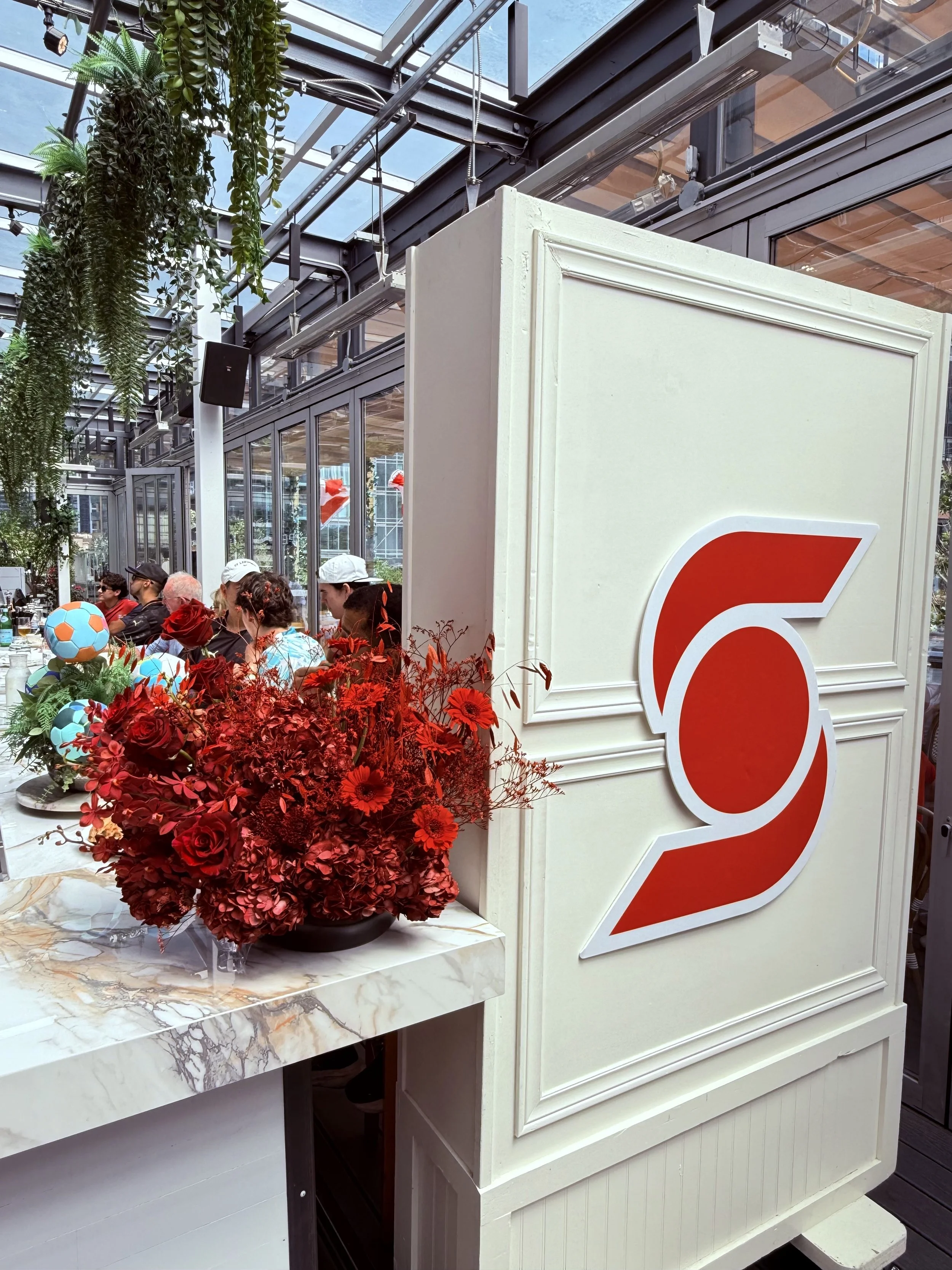 A crowded event space with people sitting at tables, a large white structure with a red number '2' on it, and hanging plants against a glass ceiling.