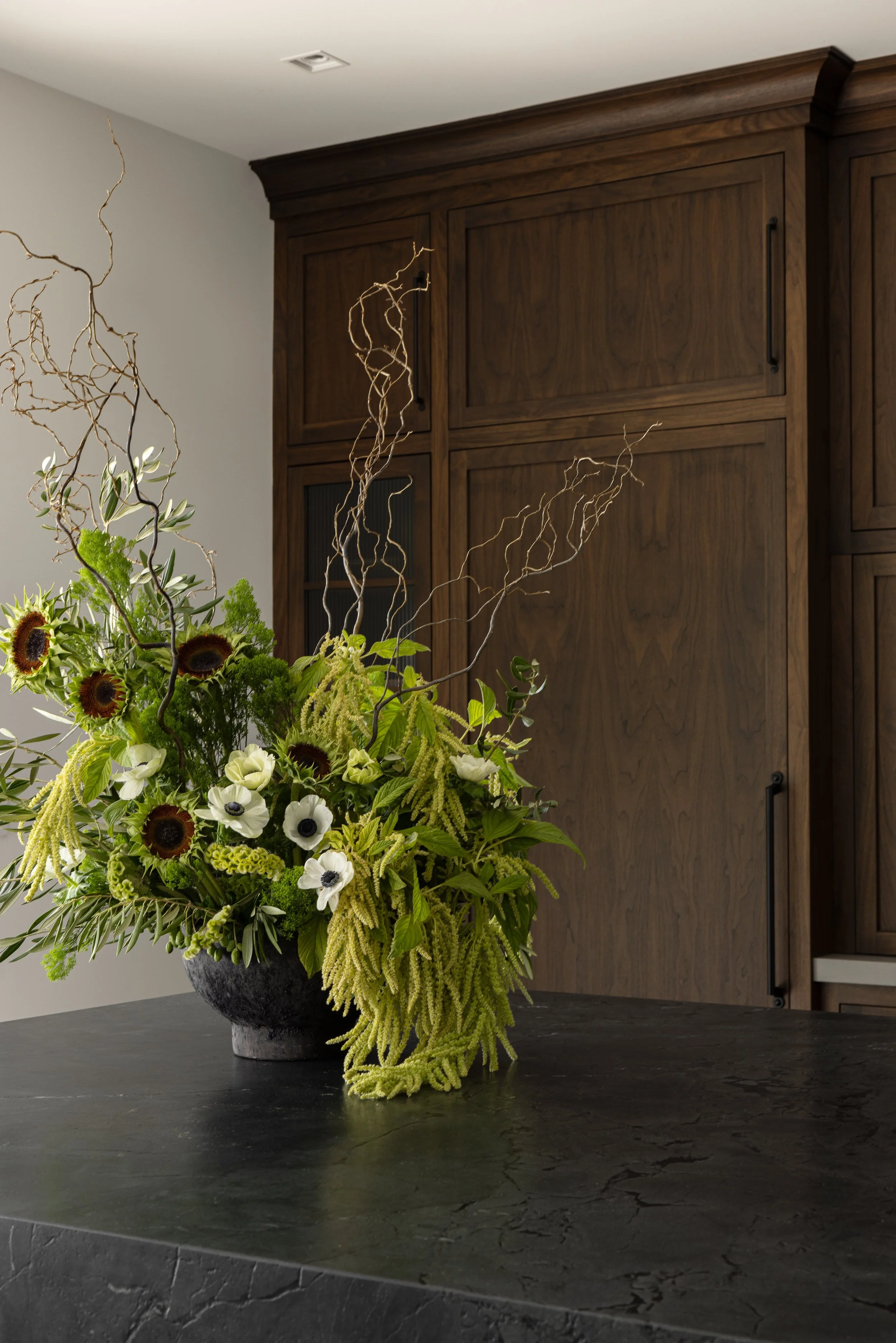 A large floral arrangement with sunflowers, white flowers, green foliage, and twisted branches in a dark gray pot, placed on a dark stone surface in front of a wooden cabinet.
