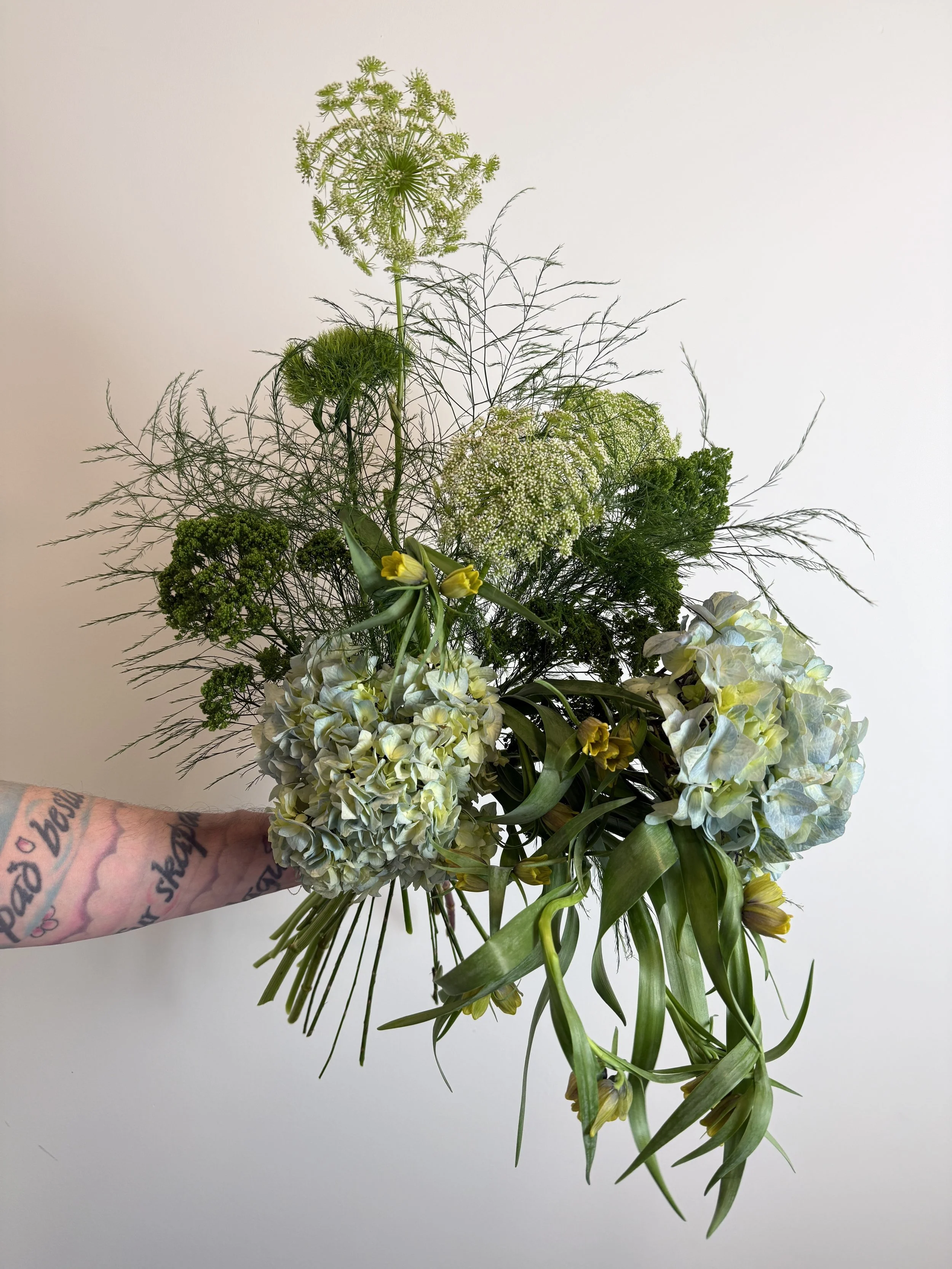 A hand holding a bouquet of various white, yellow, and green flowers and greenery against a plain white background.