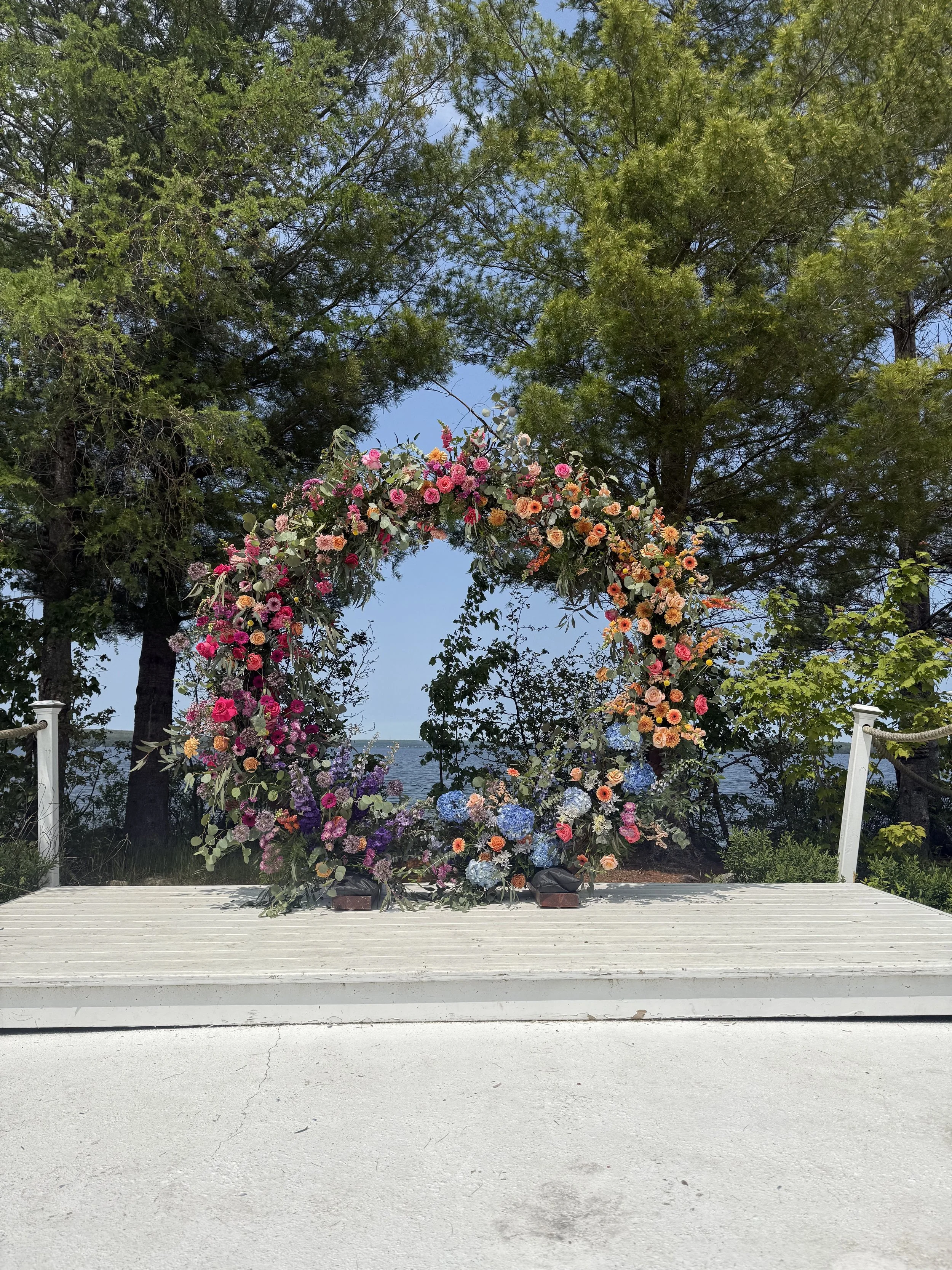A floral arch decorated with pink, orange, and purple flowers, set outdoors near a lake with tall trees and blue sky in the background.