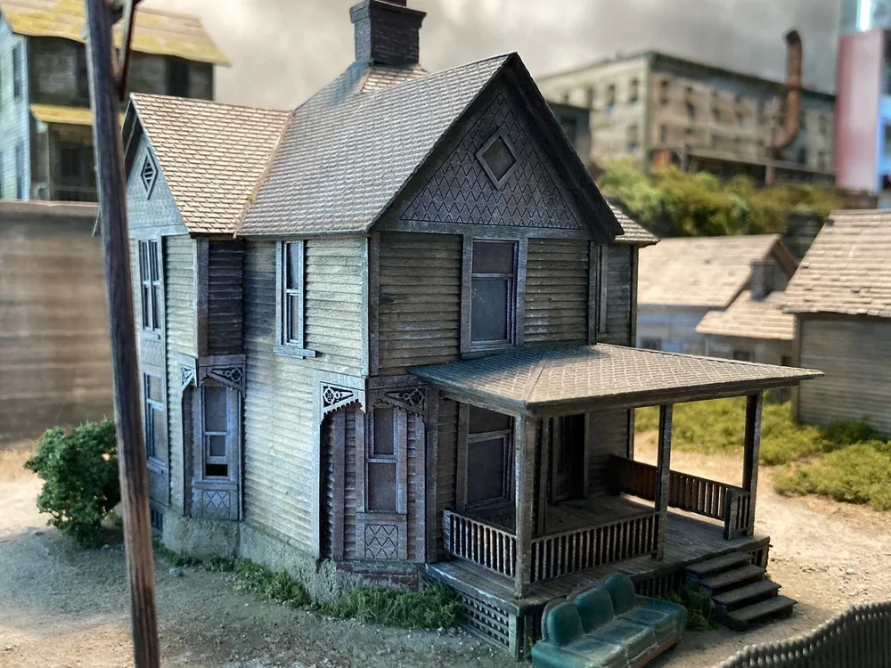 Miniature model of a Victorian-style house with a porch, steps, and detailed woodwork, placed outdoors on a sandy surface with greenery.