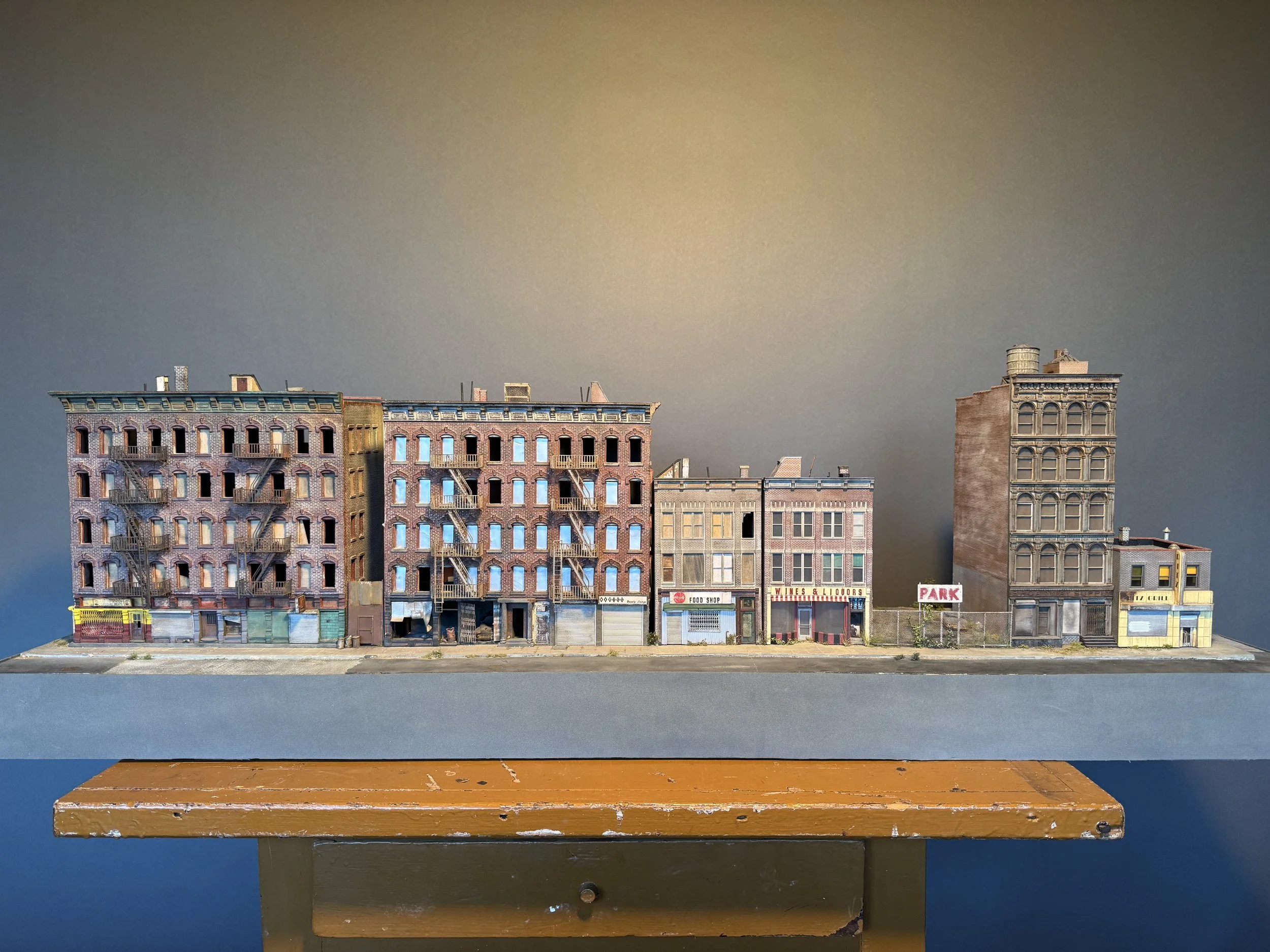 Model of a city street with colorful buildings, storefronts, and signage, placed on a platform with a wooden table in the foreground.