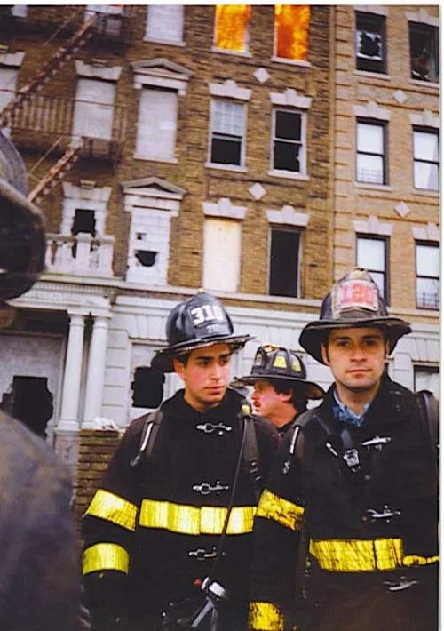 Firefighters in uniform and helmets standing in front of a burning building with flames visible through windows.