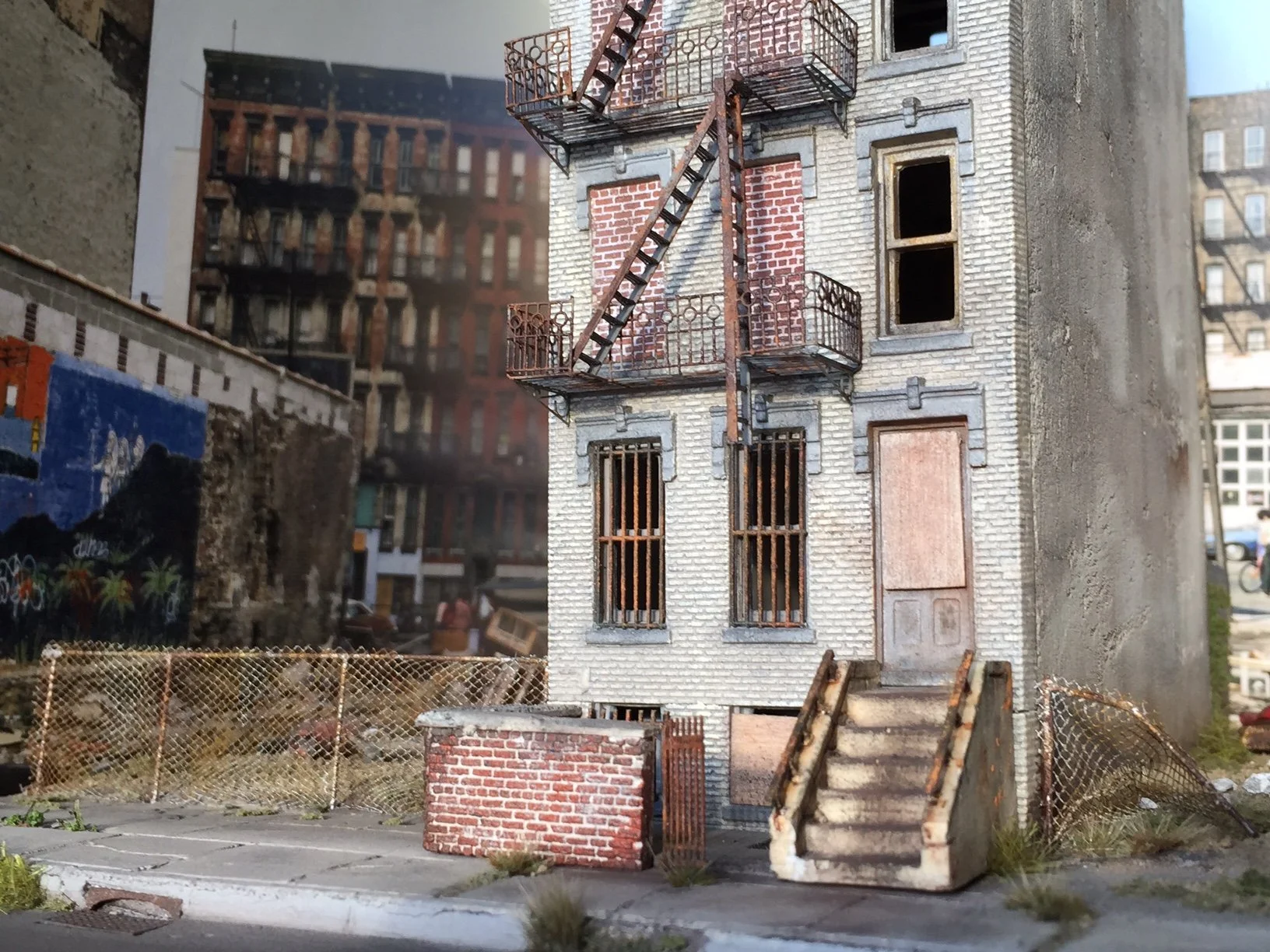 Miniature model of an old brick building with fire escapes, boarded windows, a staircase, and a fenced yard.