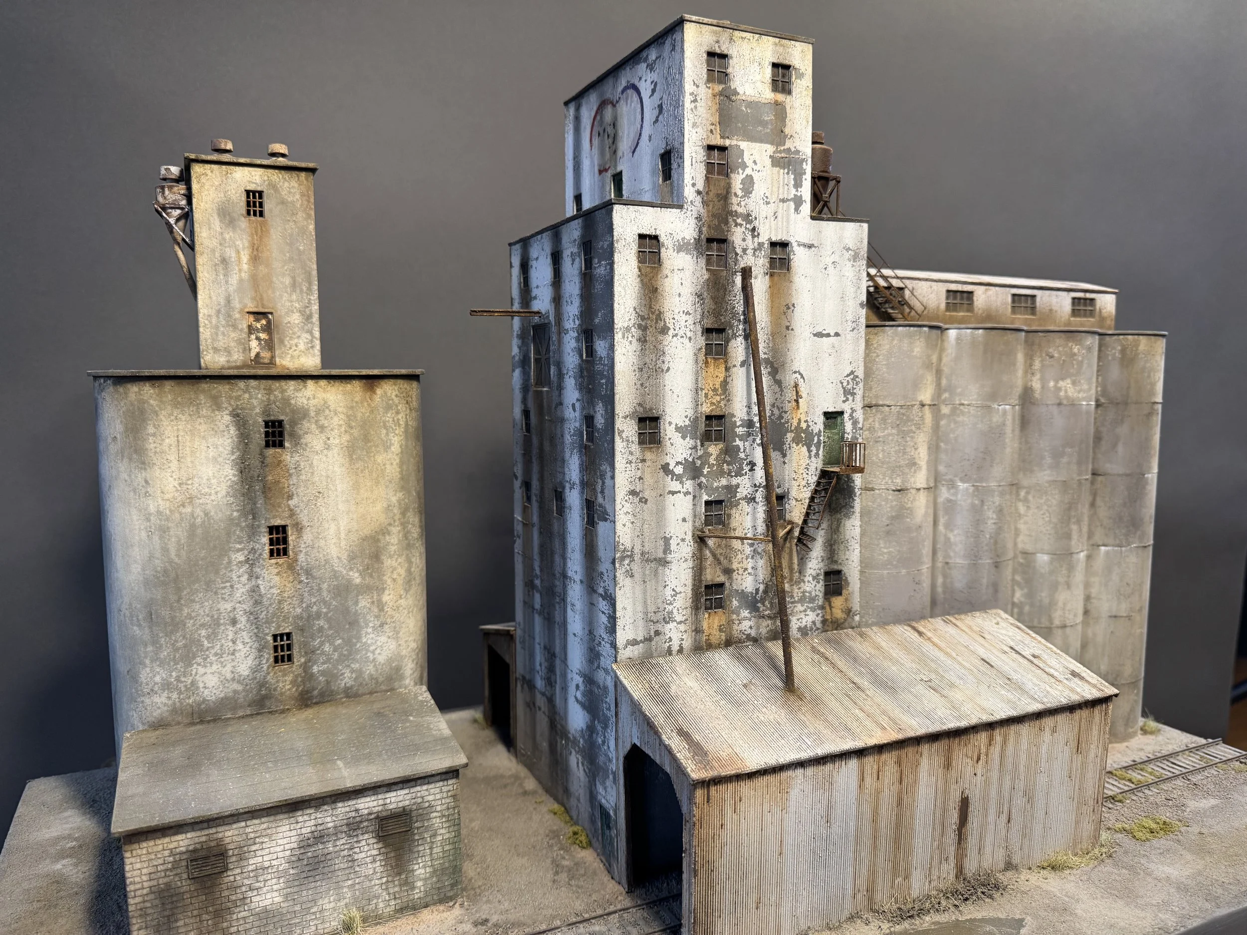 A detailed miniature model of an abandoned industrial building with weathered and rusty surfaces, small windows, external staircases, and a water tower, set against a plain gray background.