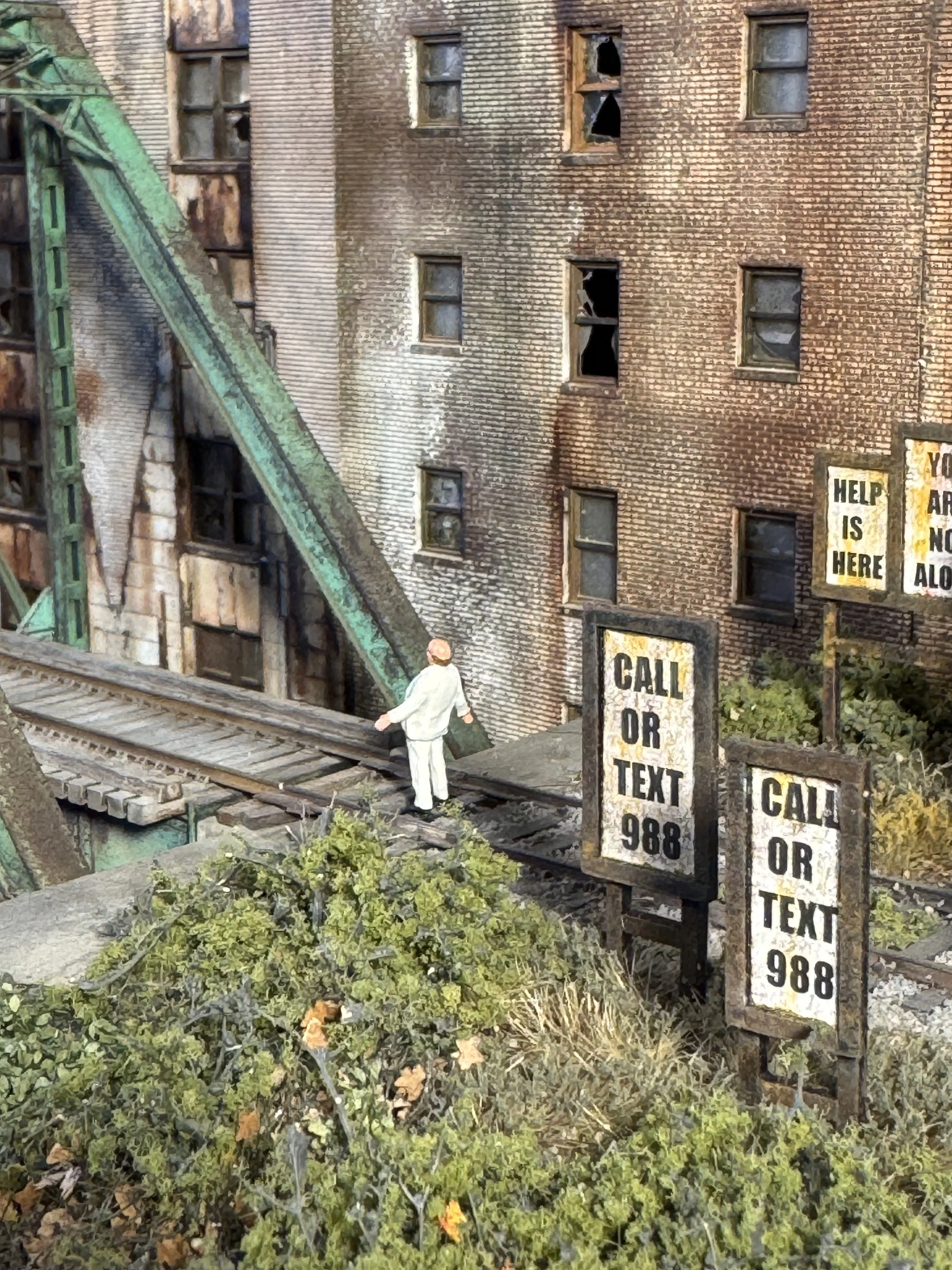 A miniature scene depicting a man in a white suit walking on a metal railway track, next to old brick buildings with broken windows, some of which have been boarded up or are missing glass. There are signs with text such as 'HELP IS HERE' and 'CALL O