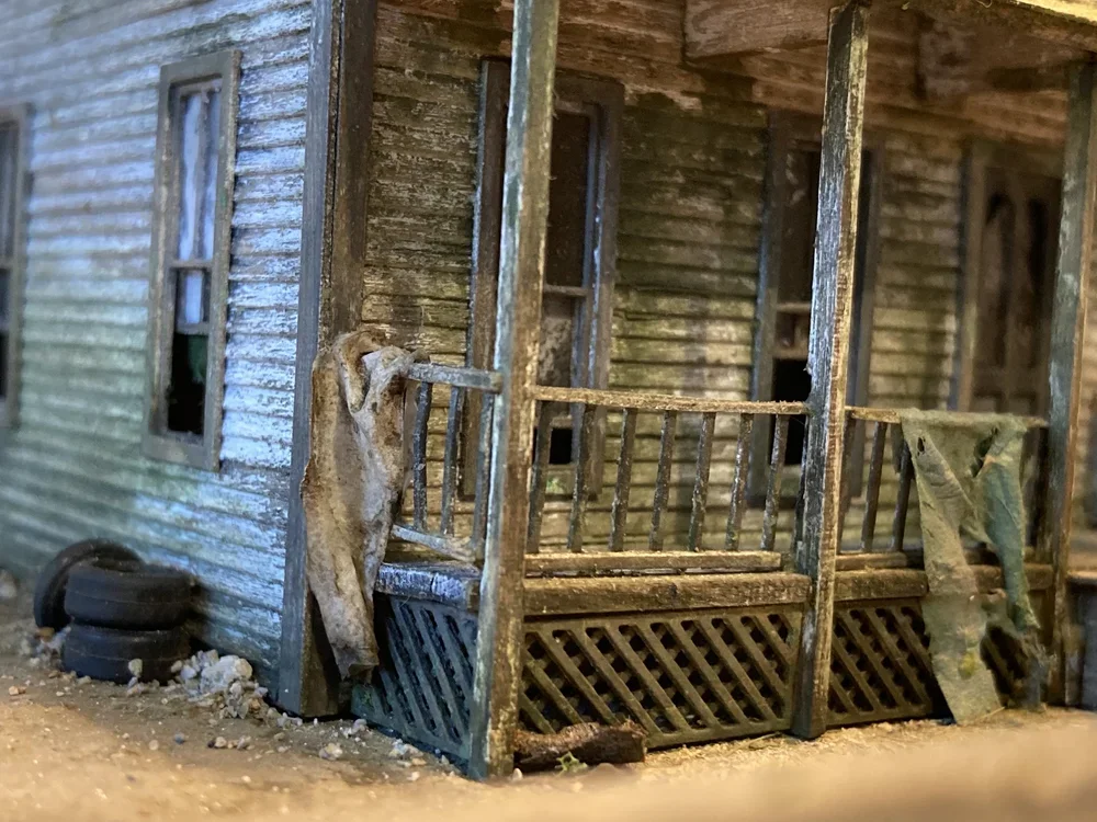 Close-up of a weathered miniature model of an old wooden house with a porch, green siding, small window frames, a tire, and some ragged cloth hanging on the porch railing.