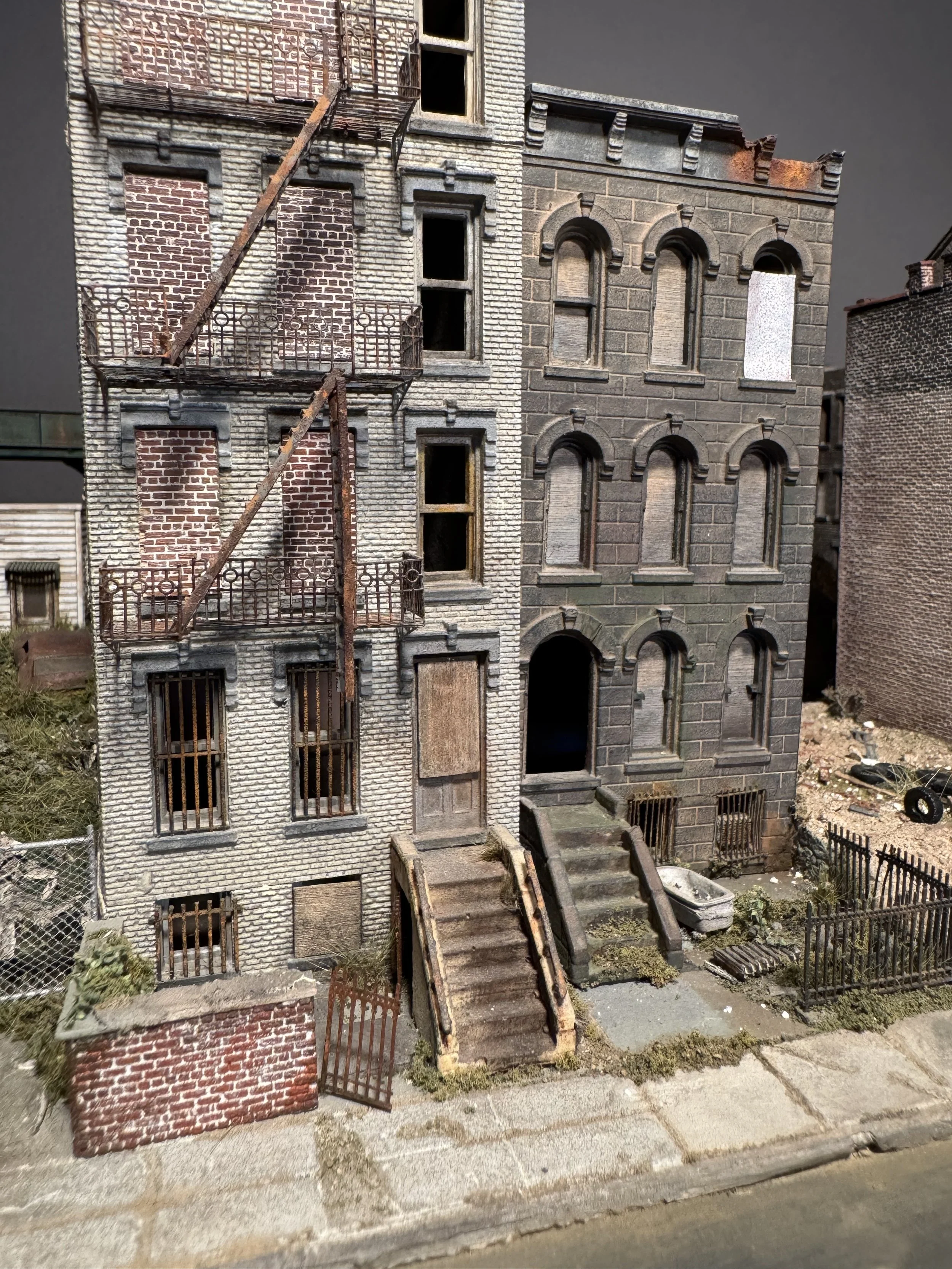 Miniature model of a dilapidated multi-story building with broken windows, rusted fire escape stairs, and overgrown surroundings.