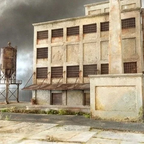 Model of an abandoned industrial building with rusted windows and a water tower in the background.