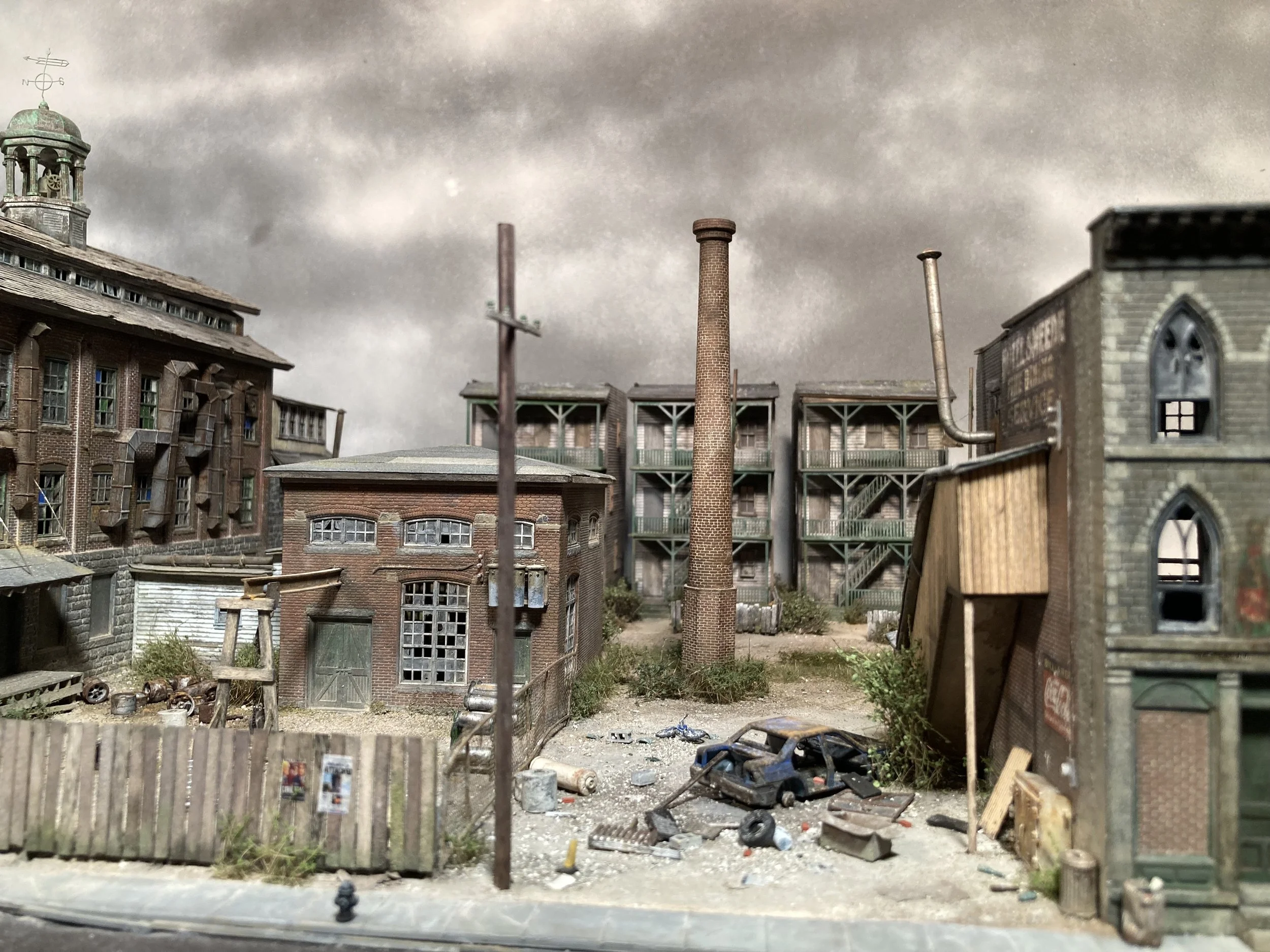 A miniature model of a dilapidated urban scene with old brick buildings, smokestacks, a wooden fence, and scattered debris.