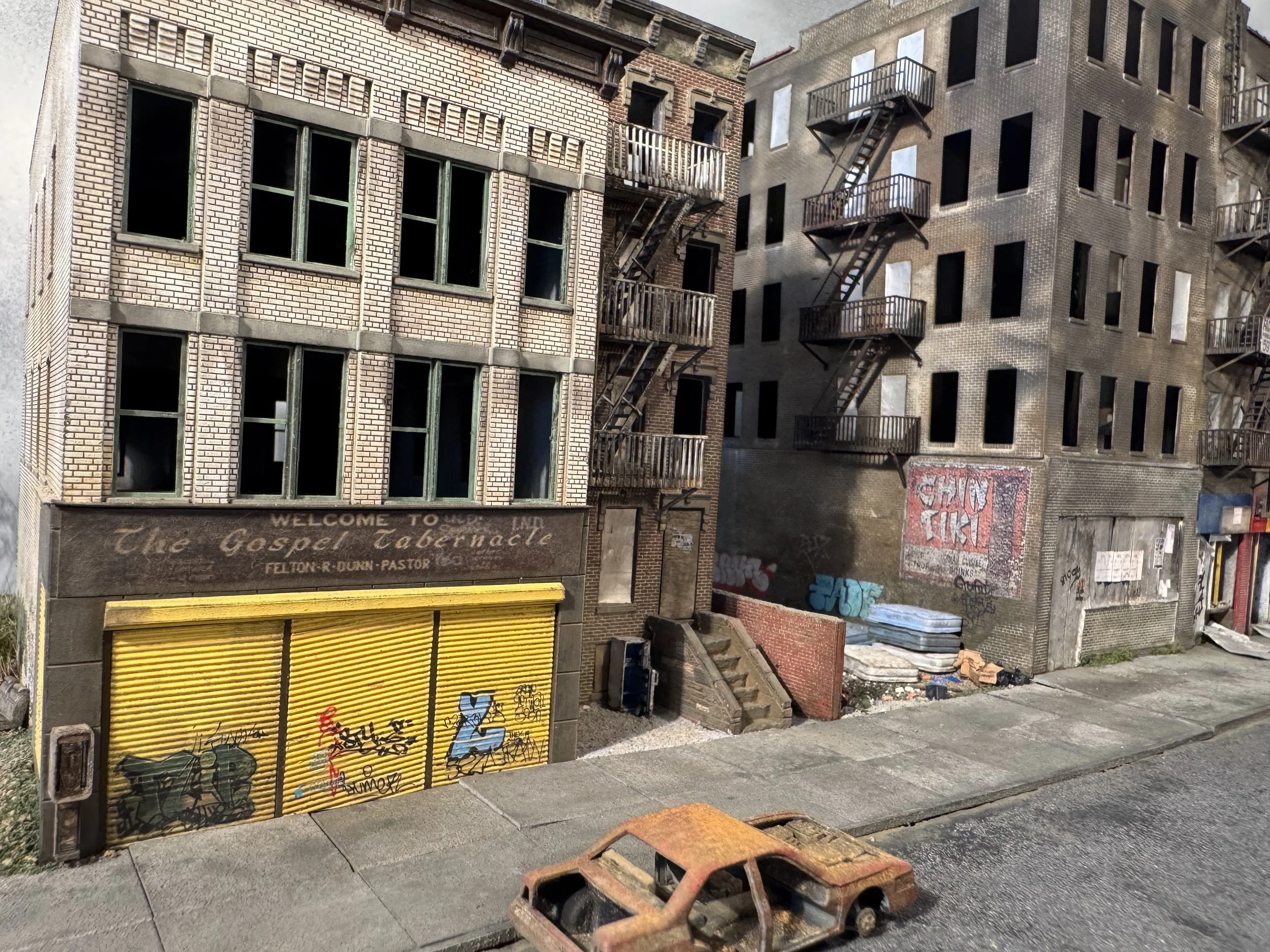 View of brick buildings with fire escape stairs, graffiti on shutters, and a rusted toy car on the sidewalk.